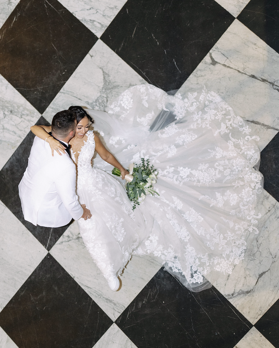 Top-down view of a bride and groom lying on a black and white checkered marble floor, with the bride holding a bouquet of greenery, dressed in a lace wedding gown.