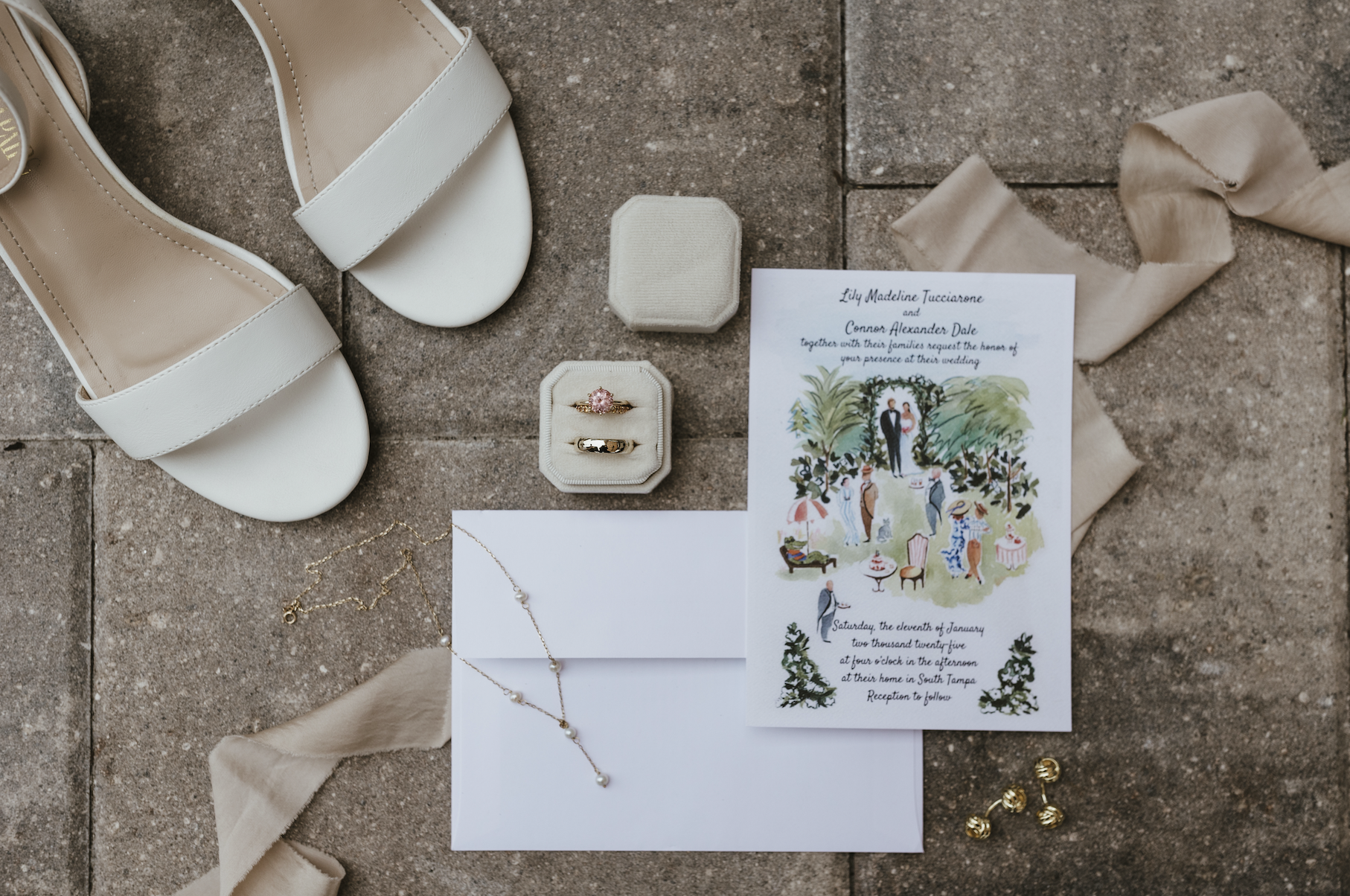 Wedding invitation, white high heel sandals, pearl necklace, gold wedding rings in a ring box, pink gemstone engagement ring, gold wedding band, beige ribbon, gold and pearl earrings, small fabric box, cream-colored cloth, and wedding invitation with floral illustration on a brick surface.