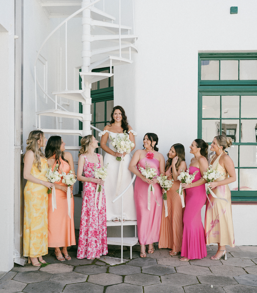 A bride in a white wedding dress standing on a spiral staircase, surrounded by seven bridesmaids in colorful dresses holding bouquets, outside near a white building with green trim.