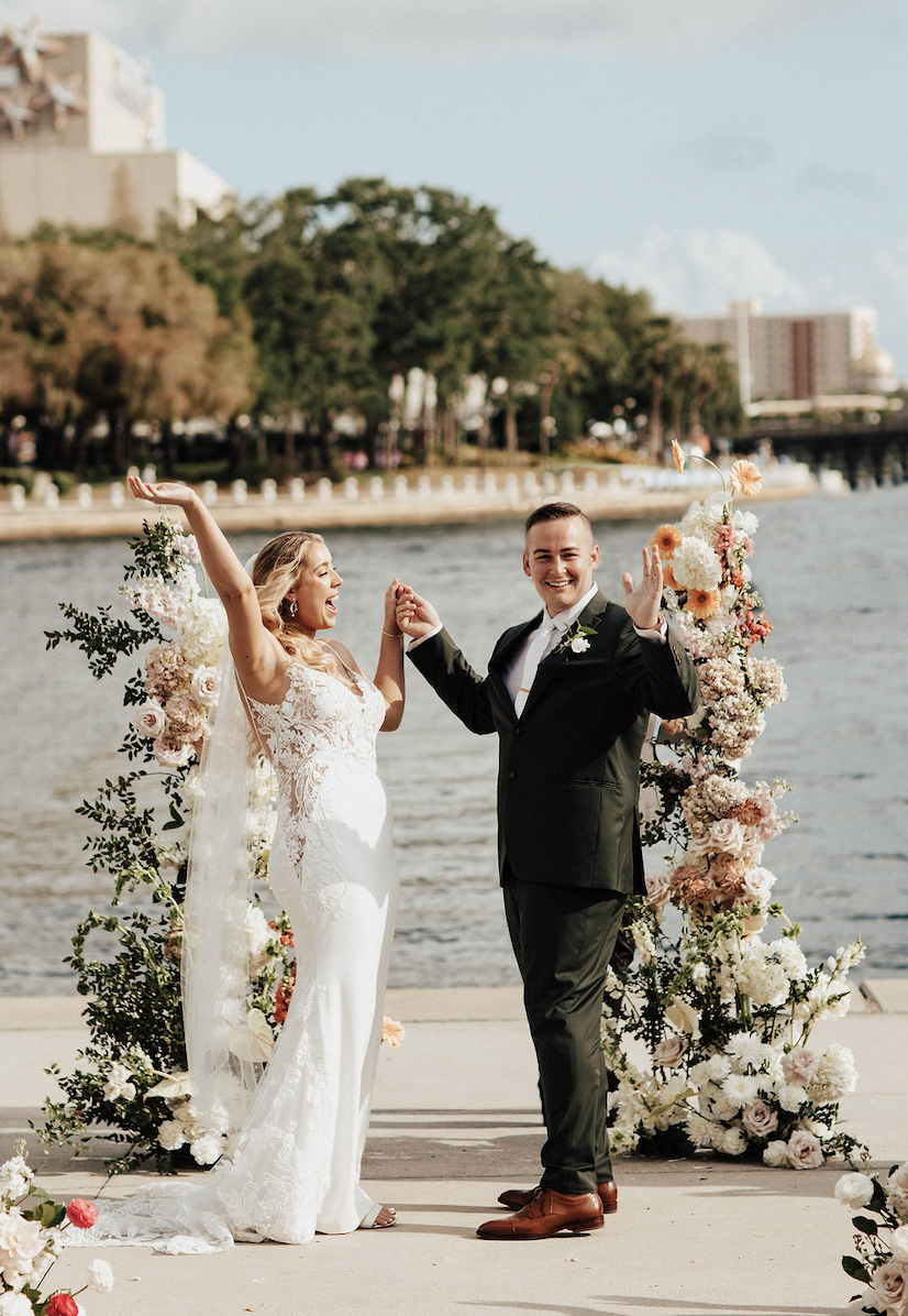 A newlywed couple celebrating their wedding by a waterfront, standing in front of a floral arch, holding hands, and smiling.