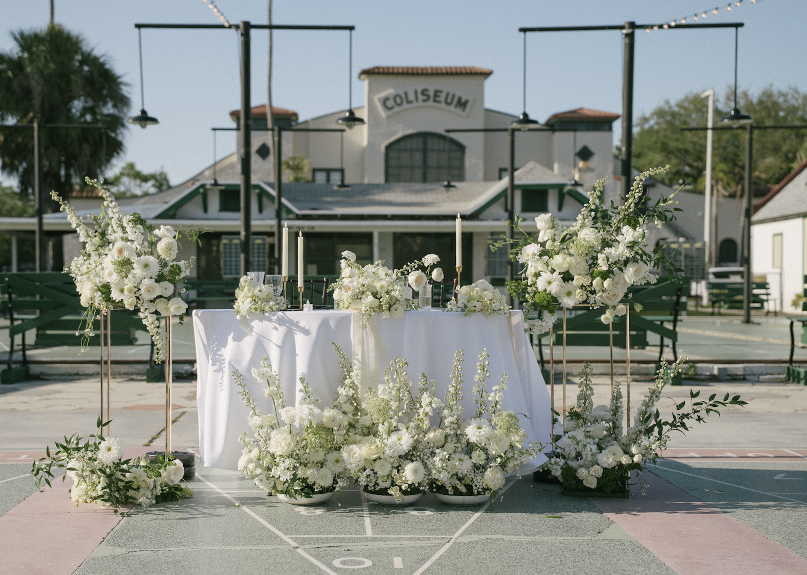 A wedding altar decorated with white flowers and candles is set up outdoors on a basketball court with a building labeled 'Coliseum' in the background.
