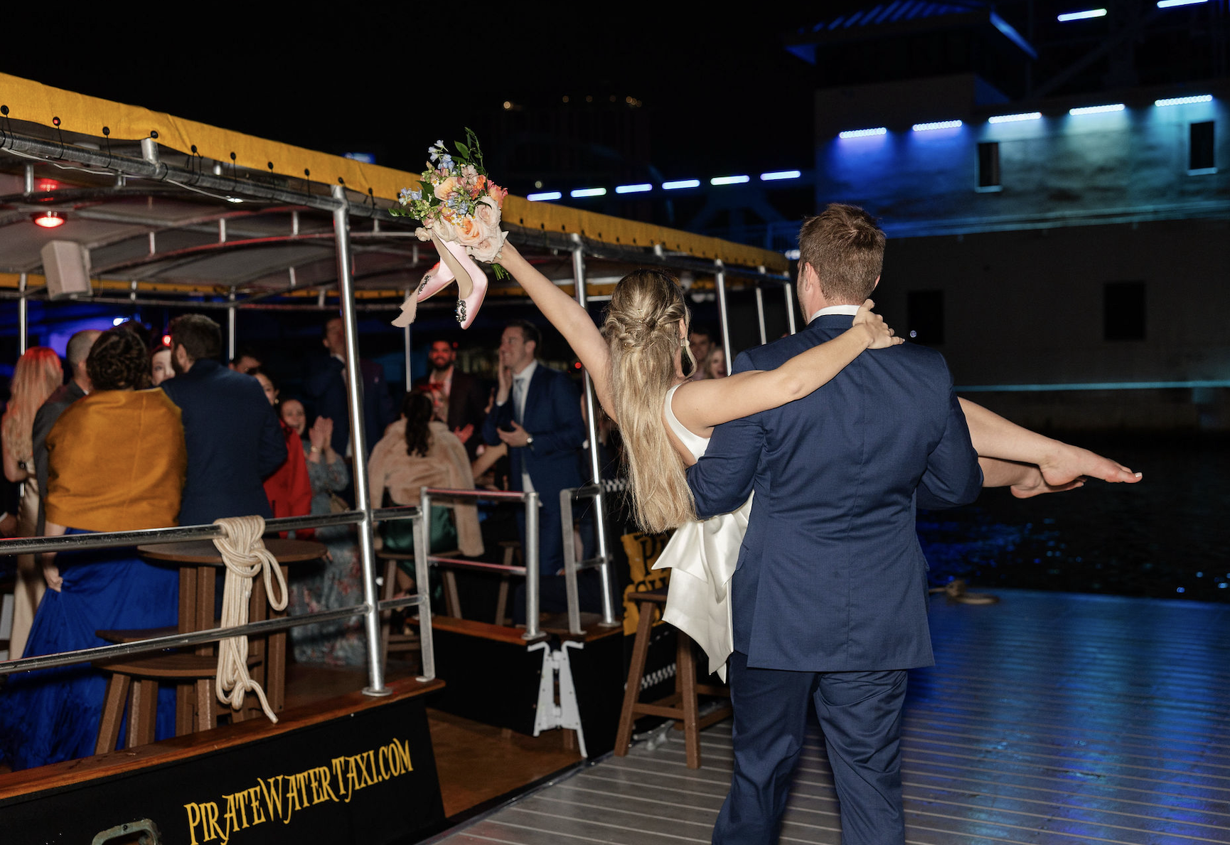 A newlywed couple dancing on a boat at night, with the bride holding a bouquet of flowers aloft and the groom lifting her. Wedding guests are visible in the background celebrating with colorful lighting and a cityscape in the distance.