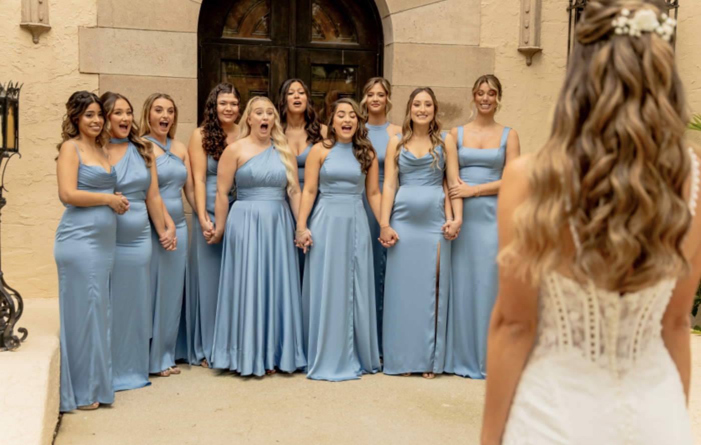 A group of ten women in matching light blue dresses are standing outside a building, looking at a woman in a white wedding dress, who is facing them.