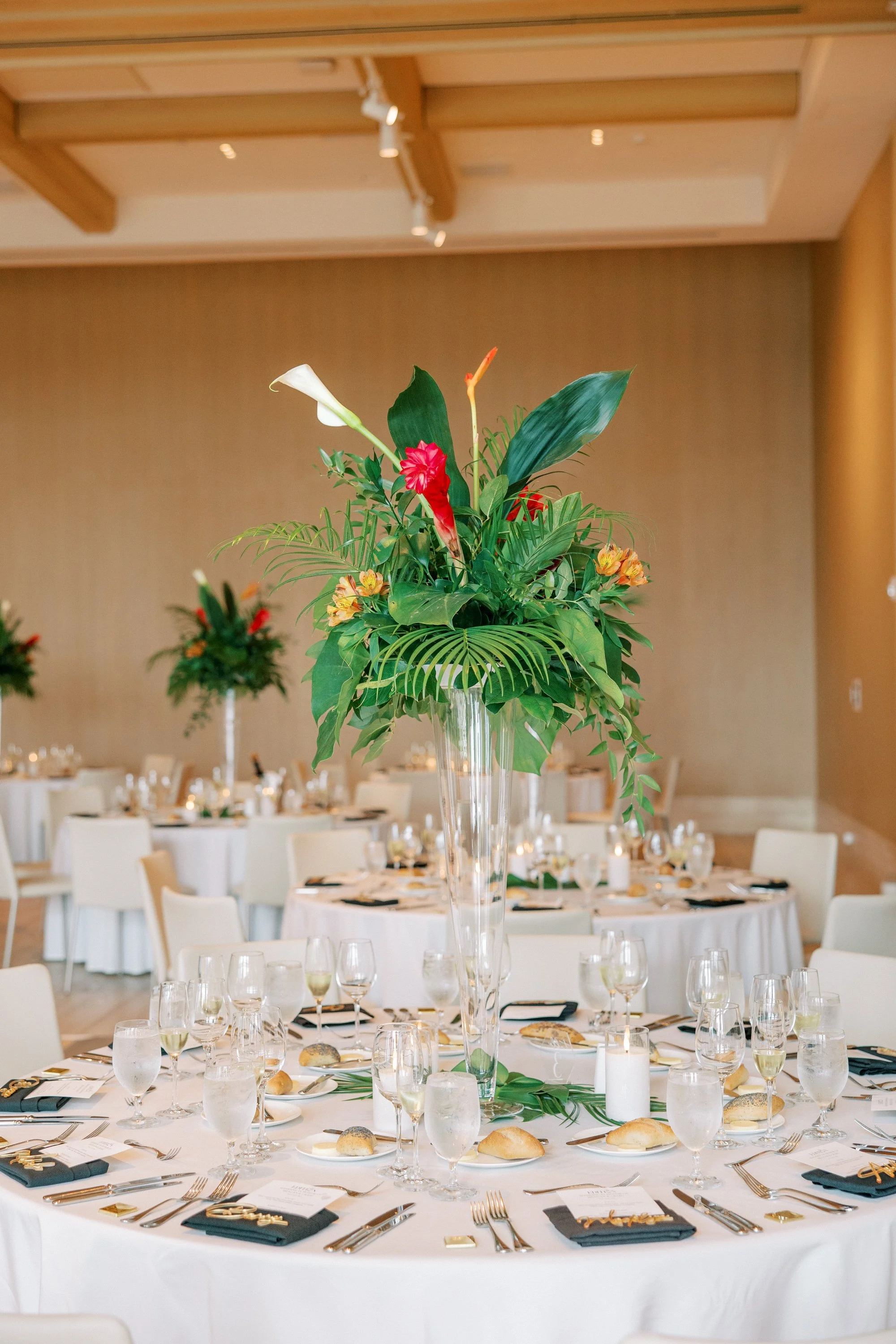 Elegant banquet table with a tall glass vase holding a lush tropical flower arrangement in a decorated event space.