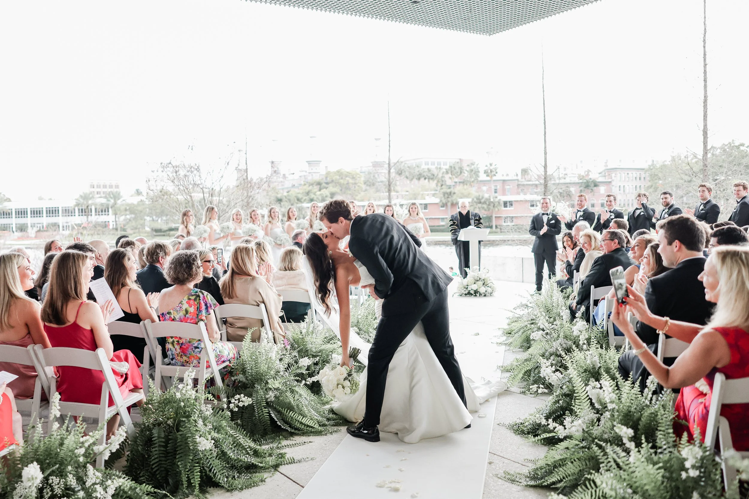 A newlywed couple kisses at their outdoor wedding ceremony under a shaded canopy, with friends and family seated on white chairs decorated with greenery, and bridesmaids and groomsmen standing in the background.