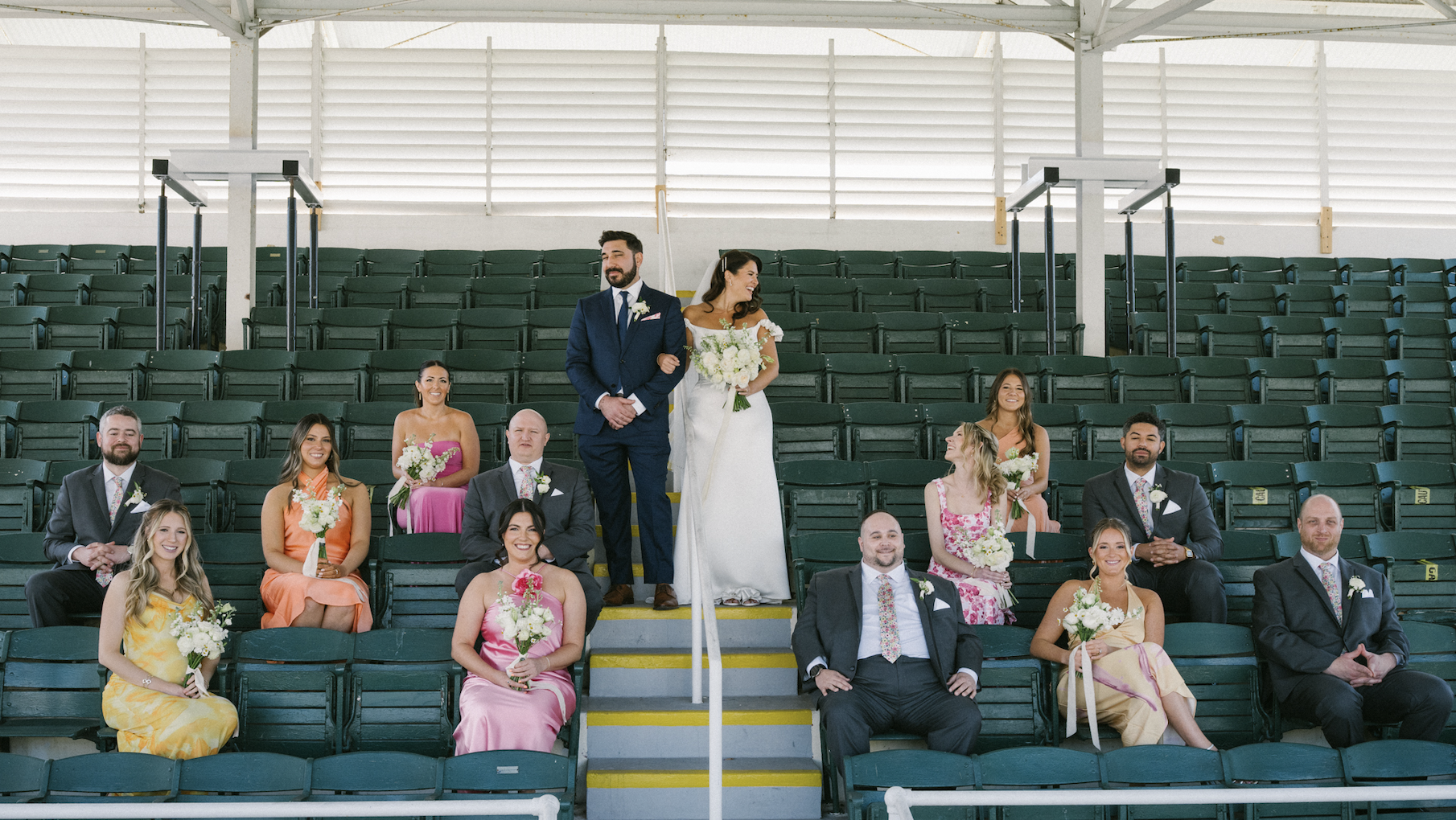 Wedding party seated in stadium seats with bride and groom standing at center, smiling and holding hands. Bridesmaids and groomsmen sitting around them, all smiling, some holding bouquets, in colorful dresses and suits.