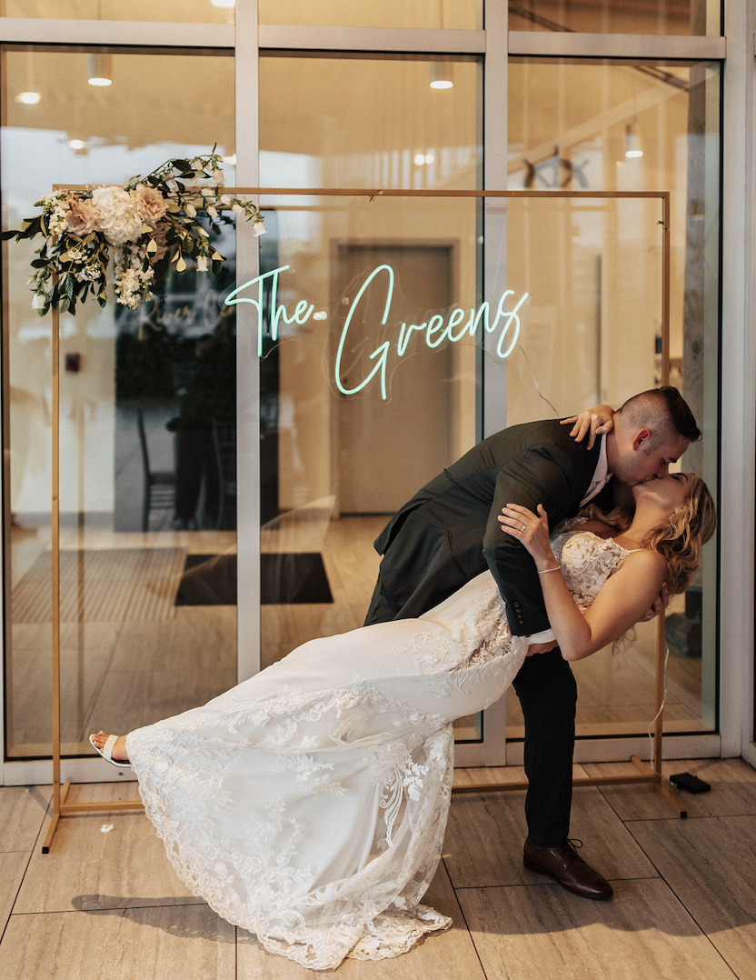 A couple in wedding attire sharing a kiss, with the groom dipping the bride, in front of a glass door with a neon sign that says 'The Greens' and floral decoration.