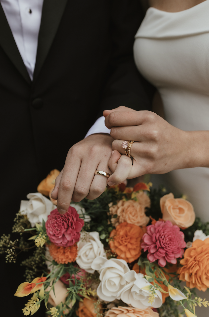 Close-up of a newly married couple holding hands with wedding rings, with a bouquet of colorful flowers in the foreground.