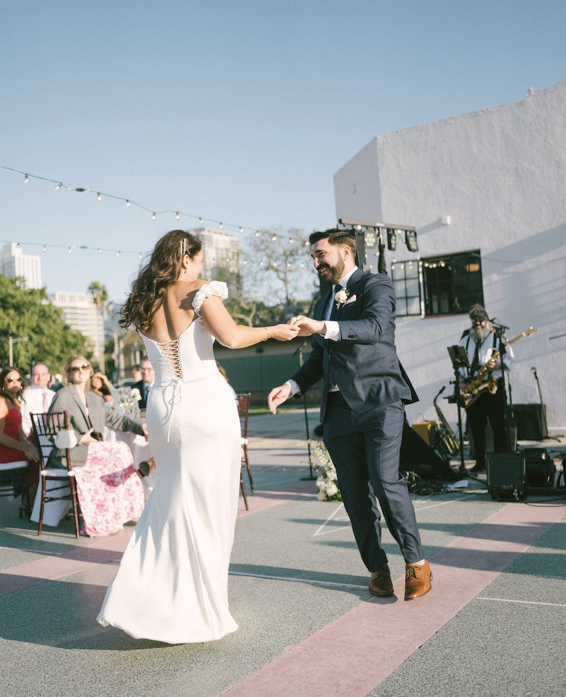 A newlywed couple dancing outside at their wedding reception with string lights, guests seated at tables, and a rock band playing in the background.