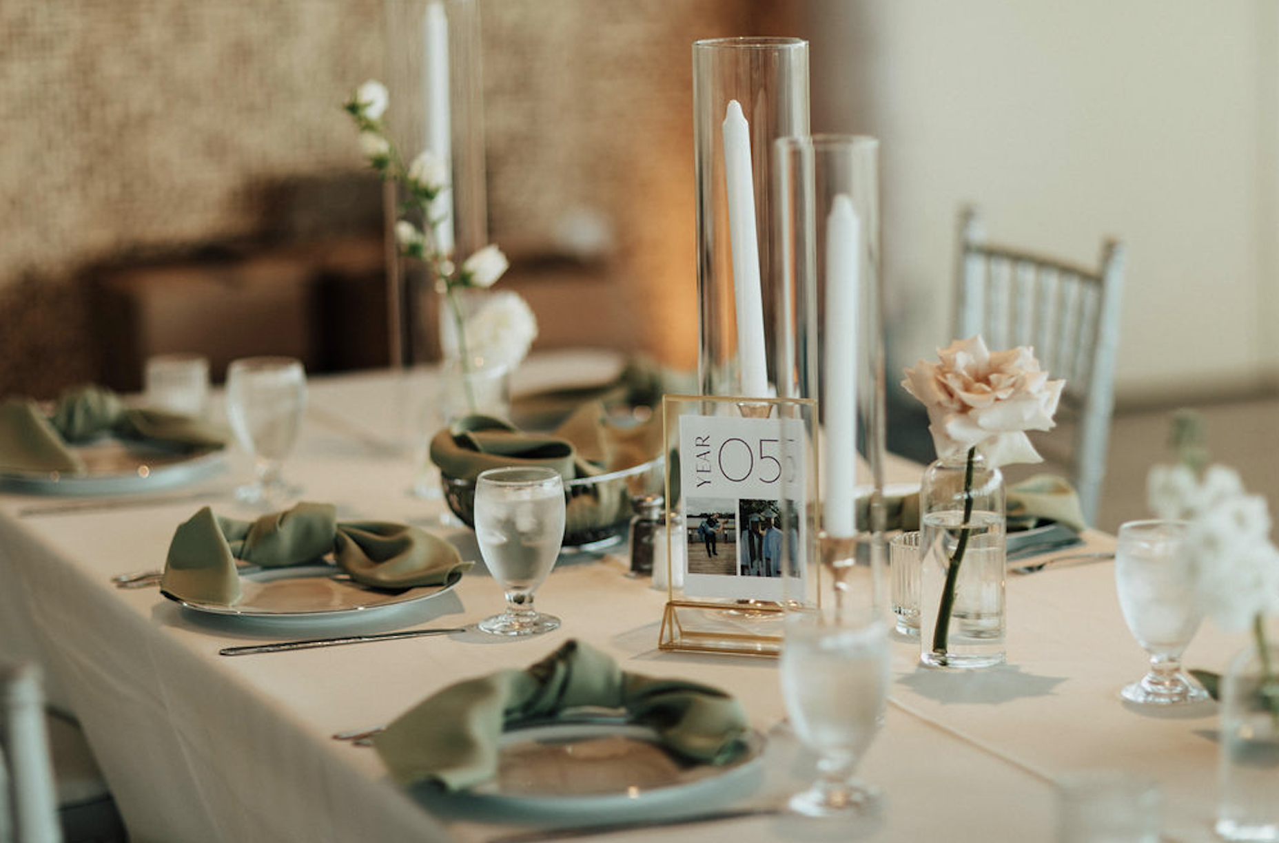 Elegant banquet table set with plates, napkins, water glasses, floral centerpieces, and tall candles, decorated in soft neutral tones for a formal event.