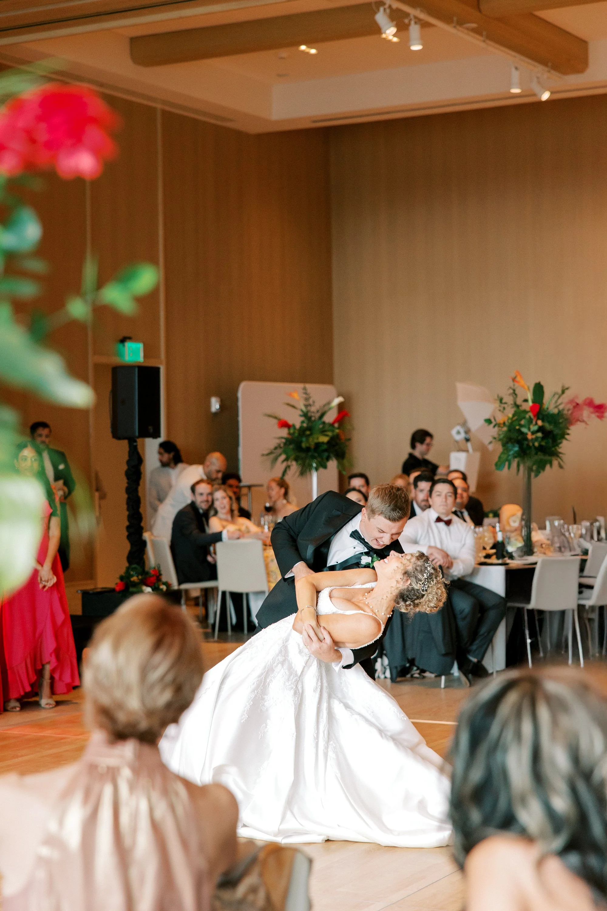 A newlywed couple dances at their wedding reception, with guests watching in the background. The groom dips the bride, who is smiling, in a ballroom decorated with tall floral arrangements.