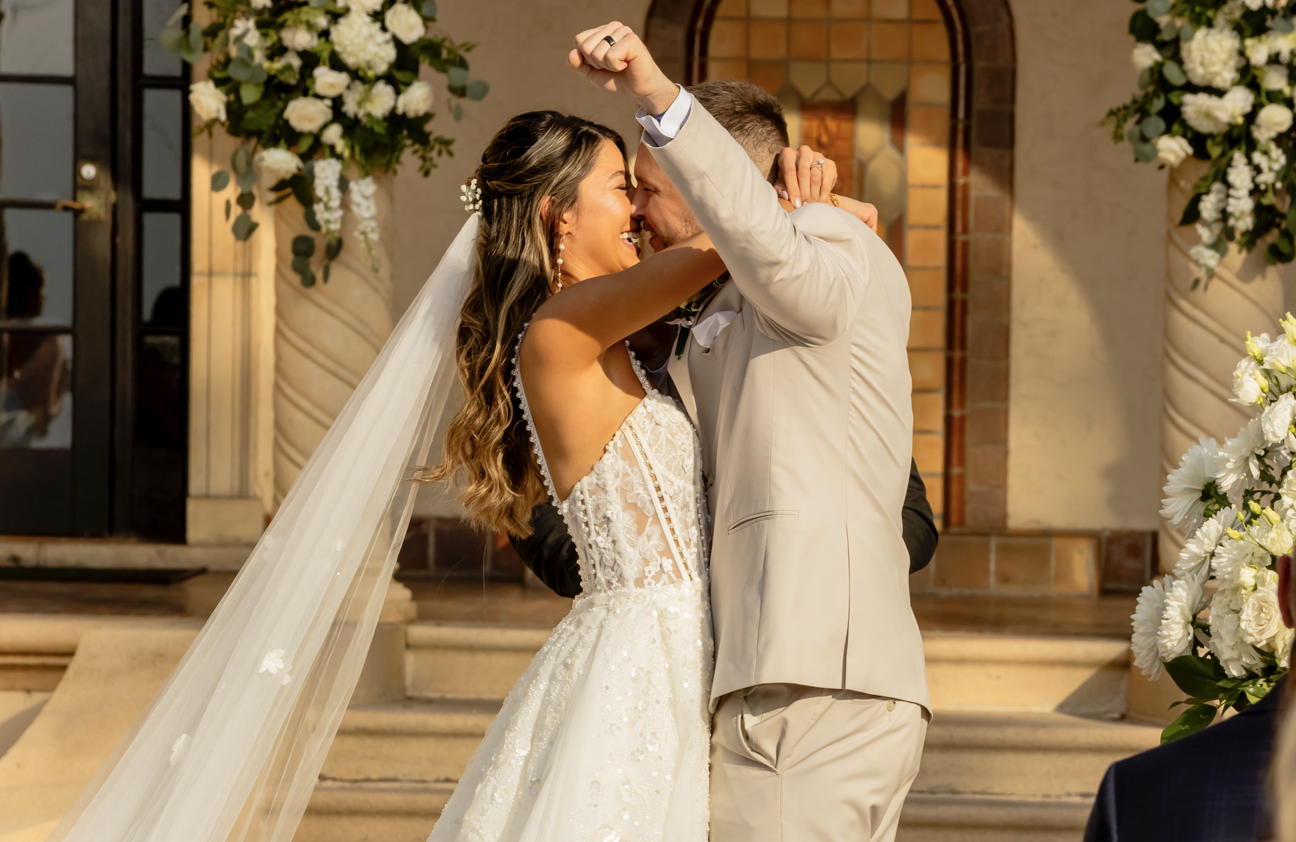 A bride and groom sharing a joyful, intimate moment during their wedding ceremony. The bride is wearing a lace wedding dress with a long veil and pearl earrings, and the groom is dressed in a beige suit. They are embracing and smiling warmly at each other on the steps outside a decorated building, with floral arrangements in the background.