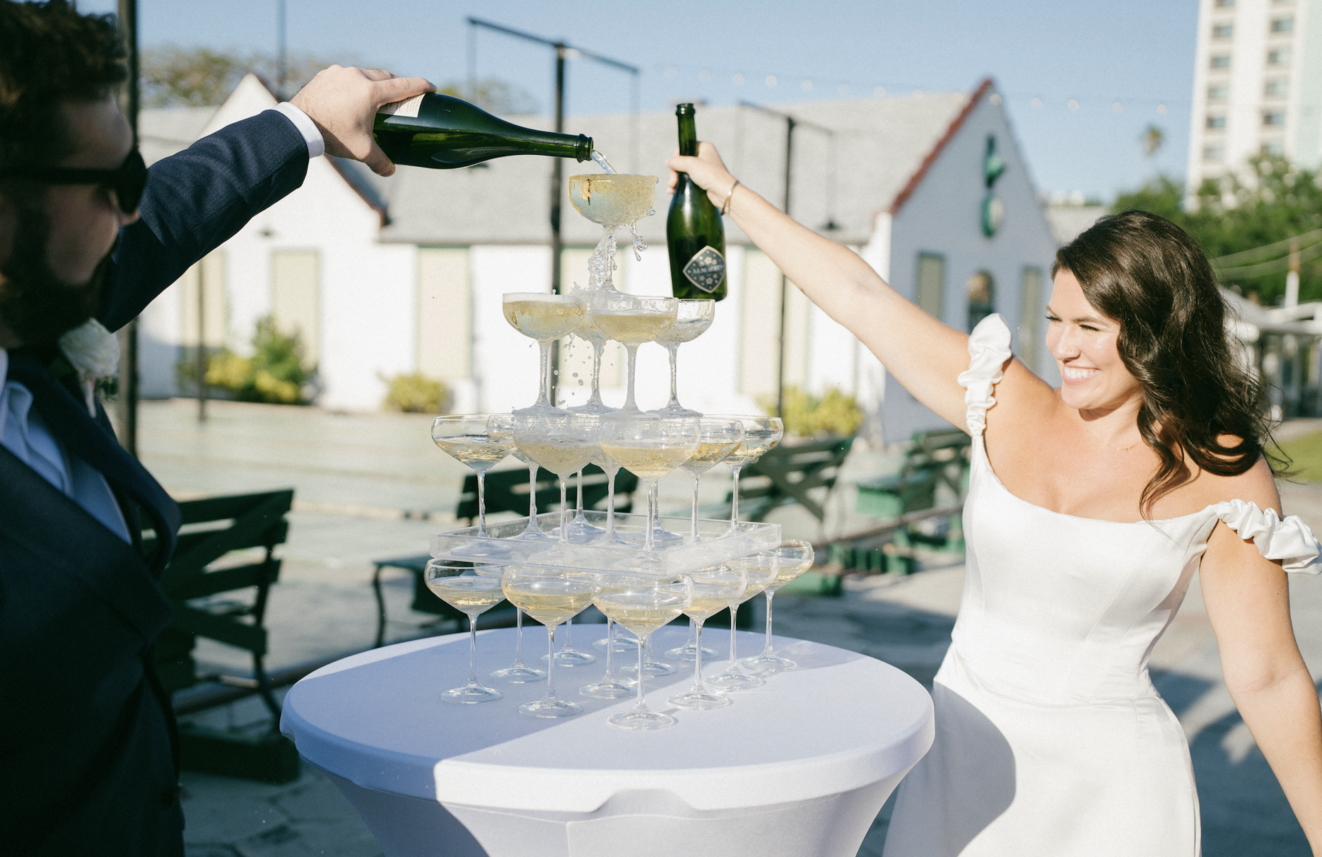 A woman in a white dress smiles as champagne is poured into a pyramid of glasses at an outdoor celebration on a sunny day.