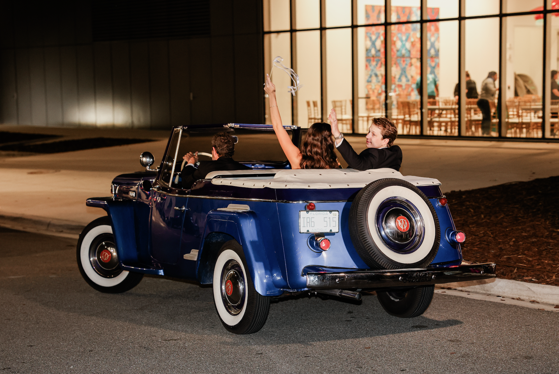 Two people riding in a vintage blue convertible car at night, celebrating and waving ribbons, with a modern building with glass walls in the background.