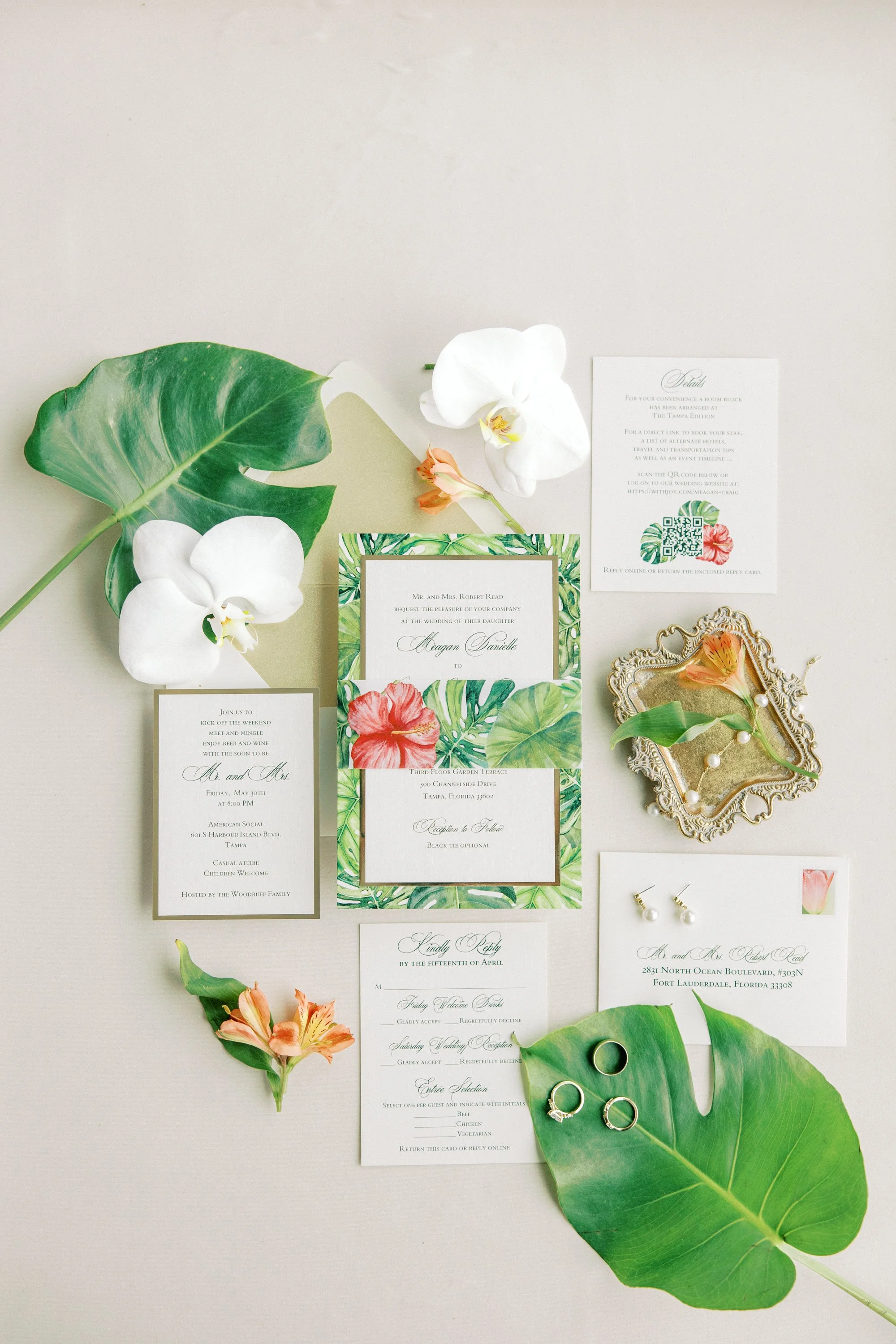 Wedding invitation suite with floral accents, greenery, and rings arranged on a white surface.