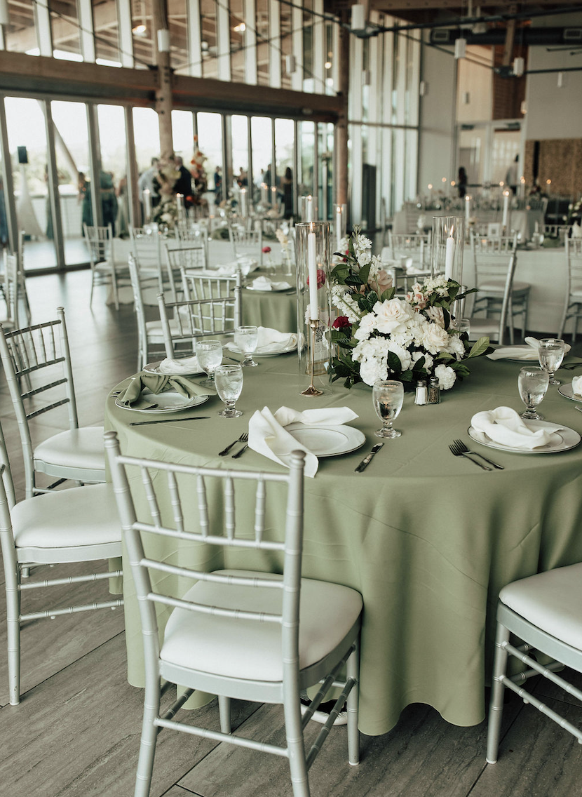 Elegant event venue with round tables covered in sage green tablecloths, set with white plates, silverware, glassware, and white napkins folded on plates. A floral centerpiece with white and pink flowers is on one table, surrounded by tall candle holders with white candles. Silver chairs surround the tables, and large windows along the walls provide natural light.