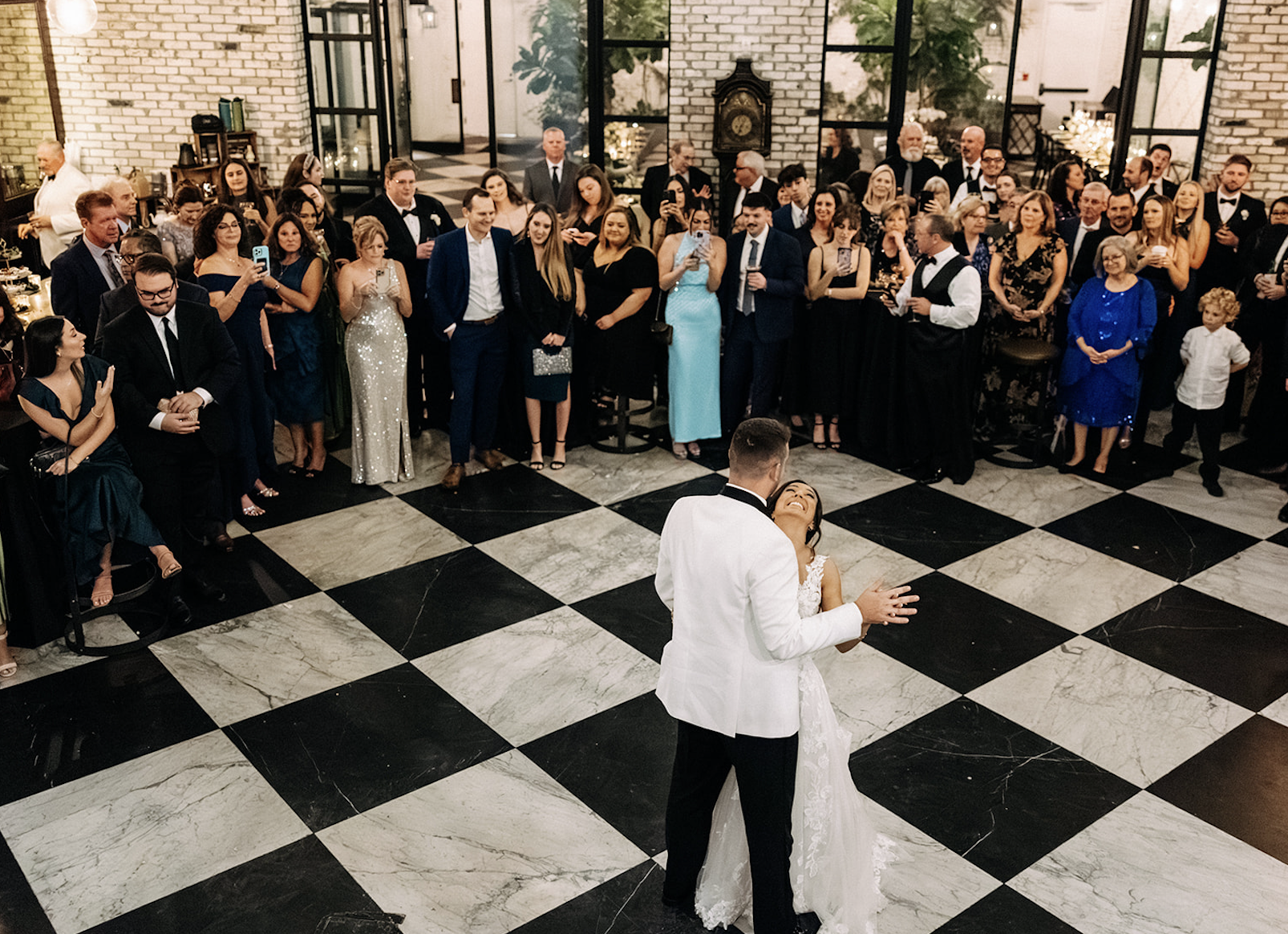 Bride and groom dancing in front of wedding guests in a ballroom with black and white checkered floor and brick walls.