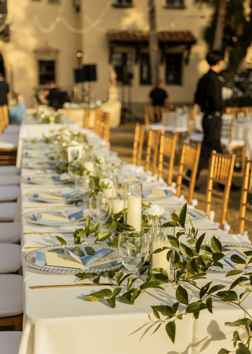 Outdoor wedding reception setup with a long table decorated with candles, green leaves, and place settings, in front of a yellow building during sunset.