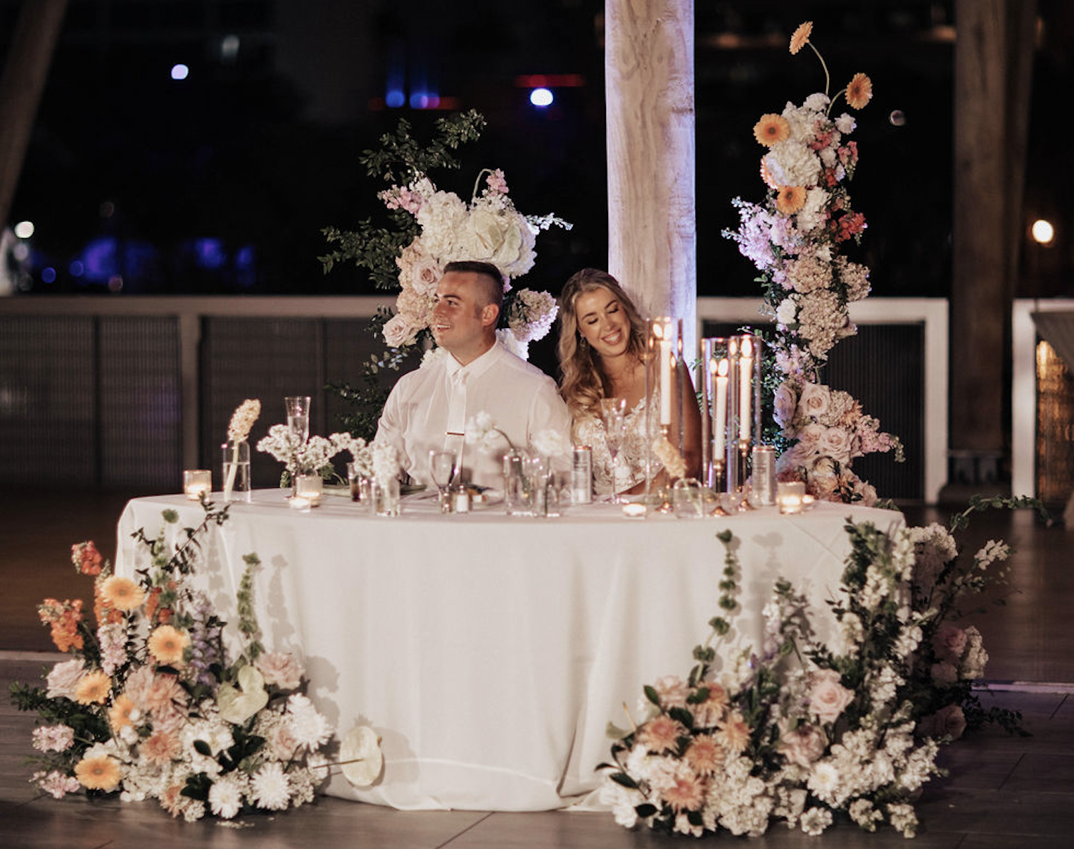 A couple sitting at a decorated wedding reception table with floral arrangements and candles.