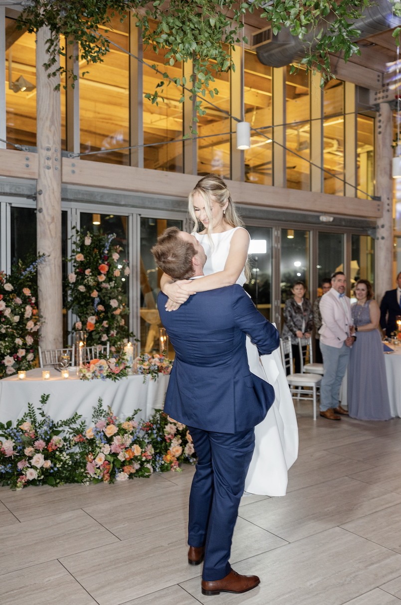 A couple dancing at a wedding reception, with the groom lifting the bride in his arms. The room is decorated with flowers and candles, and guests are watching in the background.