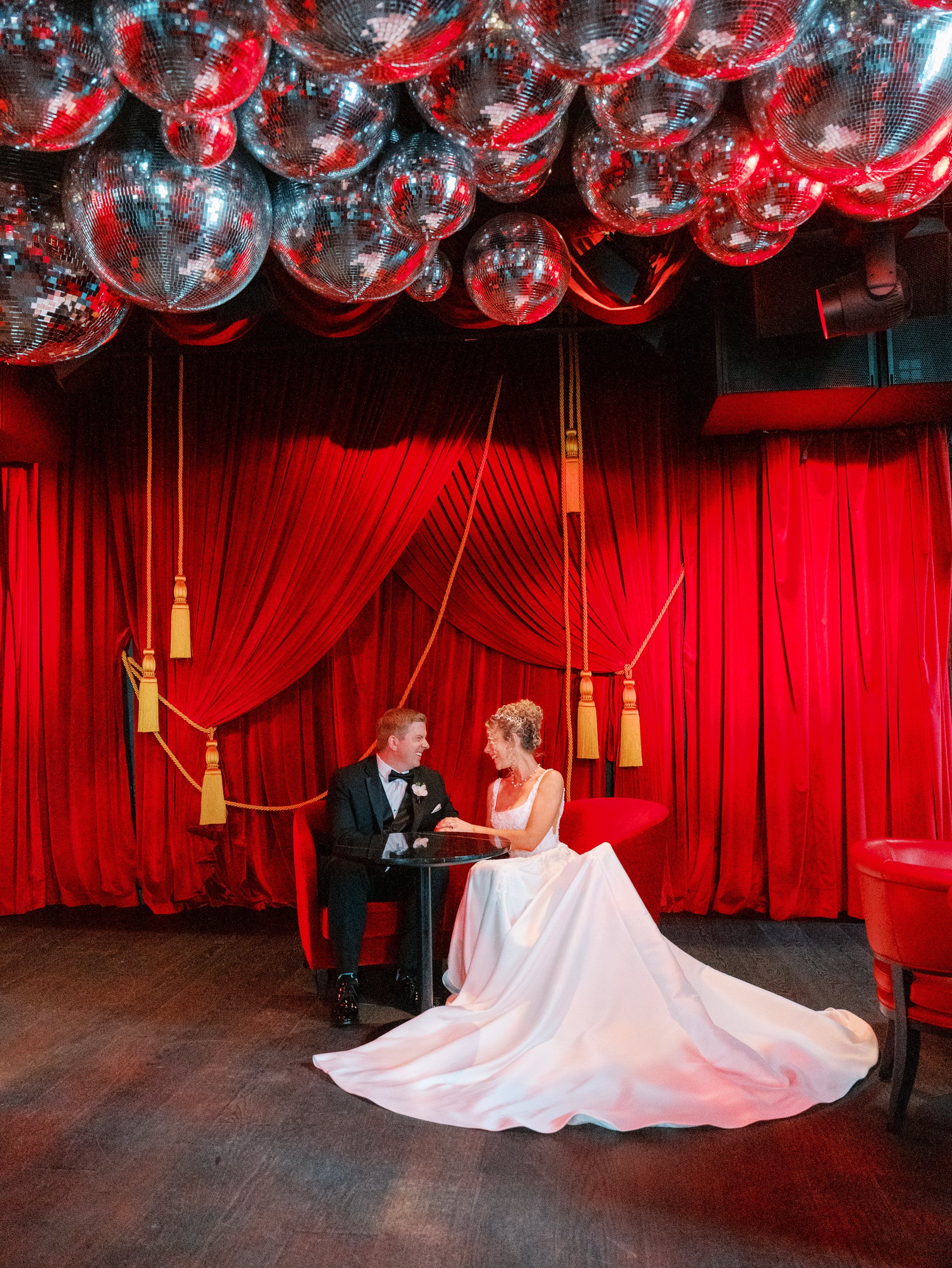 A bride and groom sitting at a small table on a stage with red velvet curtains and red hanging disco balls.