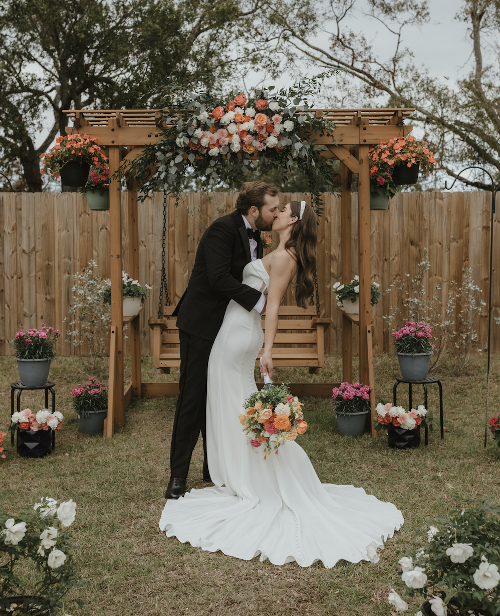 A bride and groom share a kiss during their wedding ceremony outdoors, with the bride holding a bouquet of orange and pink flowers and the groom in a tuxedo, standing in front of a decorated wooden arch with flowers and greenery.