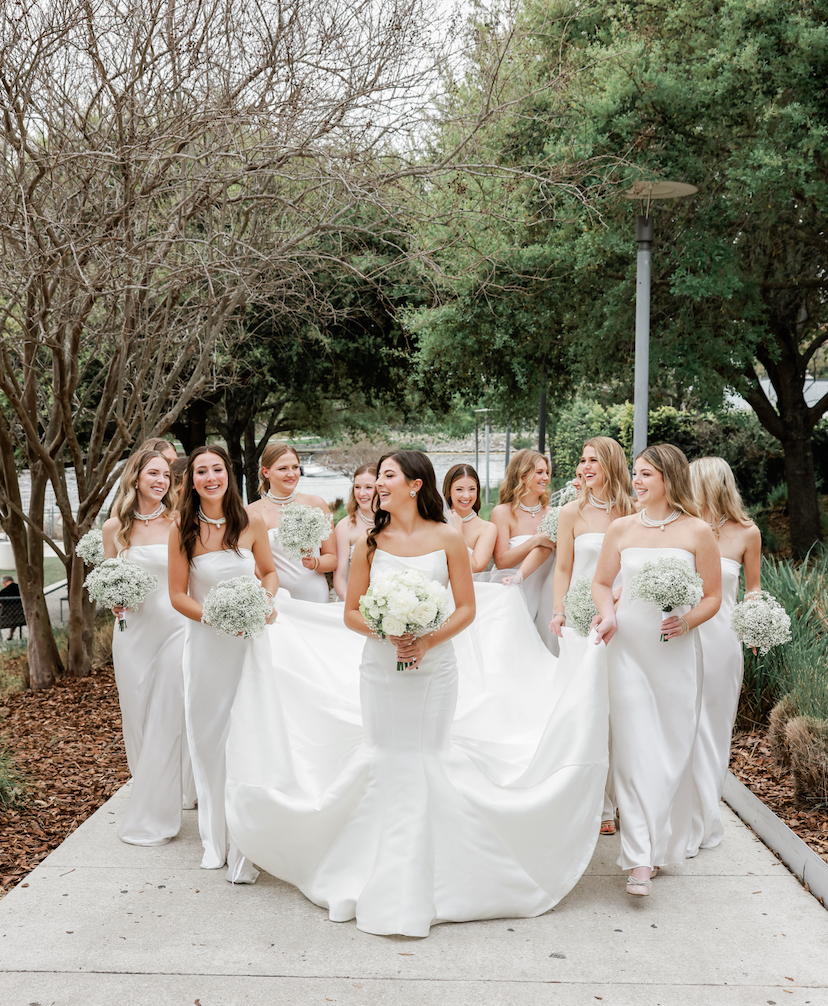 A bride and her bridesmaids in white dresses walking outdoors on a paved path surrounded by trees and bushes, holding bouquets of white flowers, smiling and laughing.