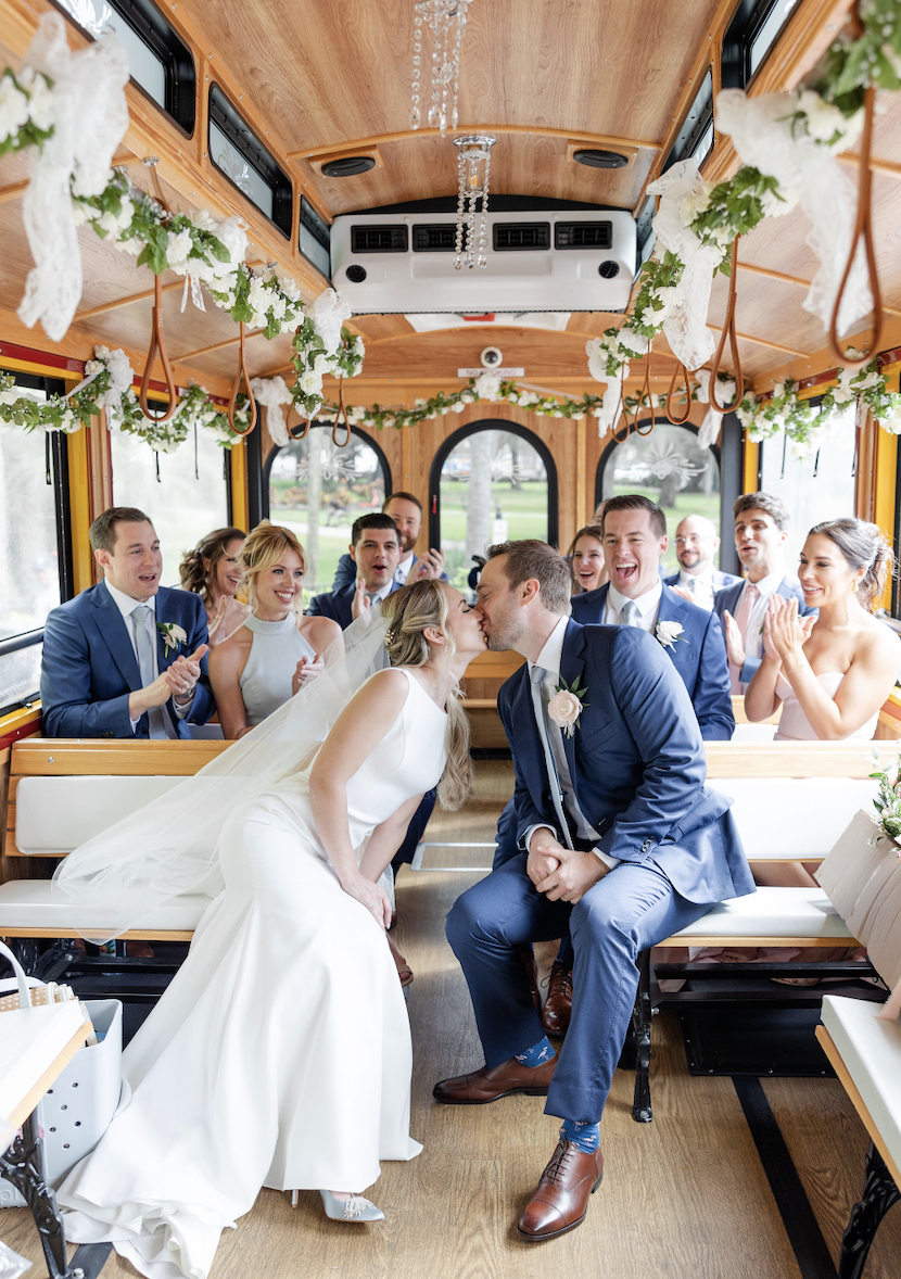 A wedding ceremony inside a decorated trolley with a bride and groom kissing, surrounded by smiling friends and family.