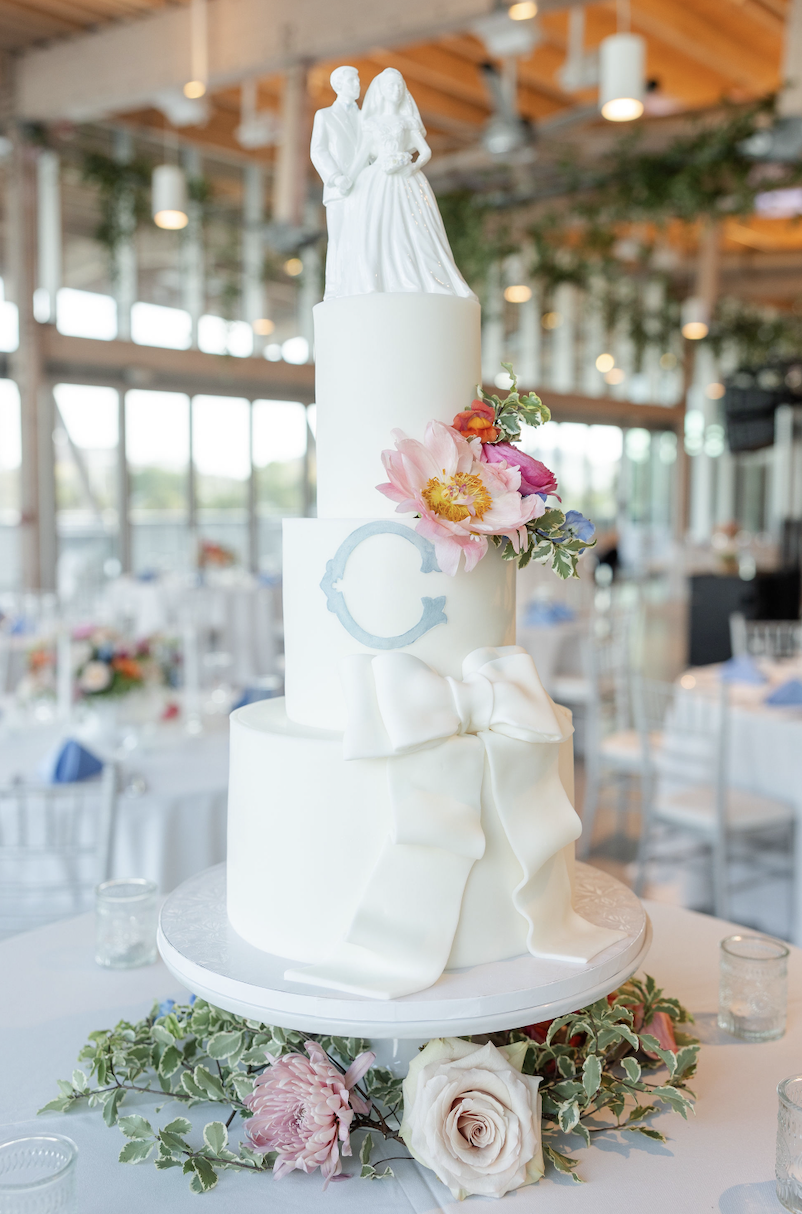 A tiered wedding cake decorated with a white fondant bow and pink flowers, topped with a figurine of a bride and groom, surrounded by greenery and flowers on a table.