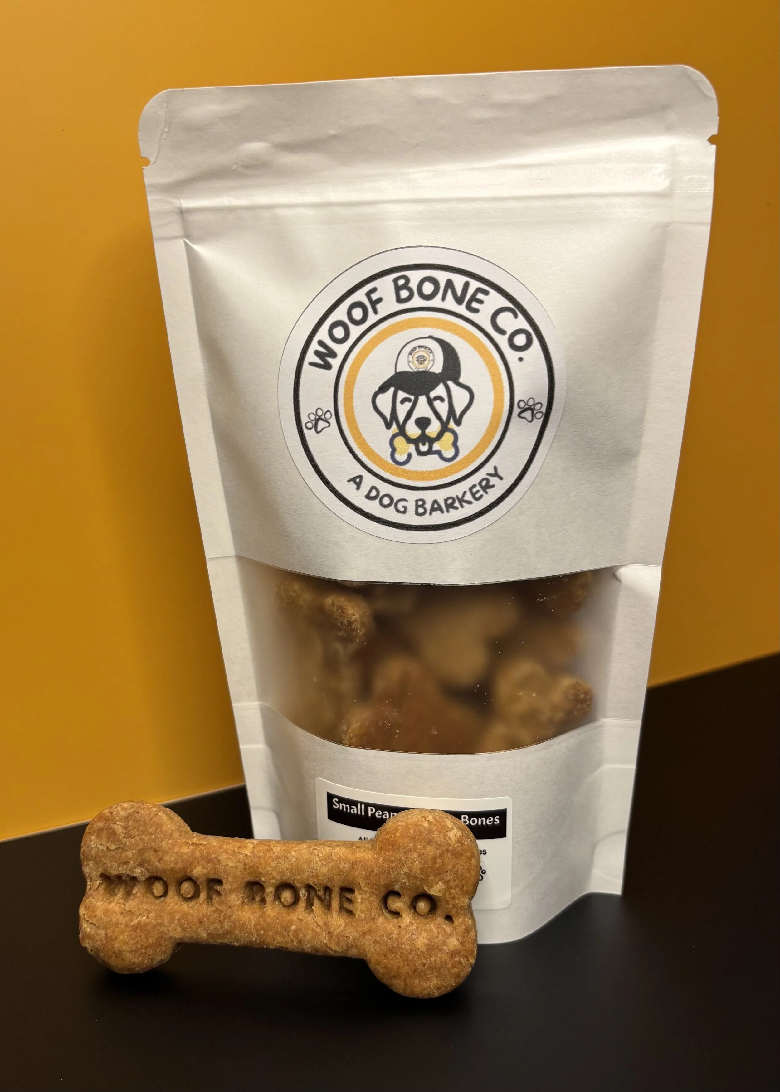A package of Woof Bone Co dog treats with a bone-shaped treat in front, set against a black surface and a yellow background.