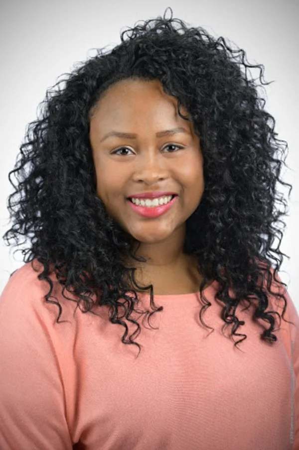 Channell, a woman with curly black hair, smiling, wearing a light pink top, against a plain light-colored background.