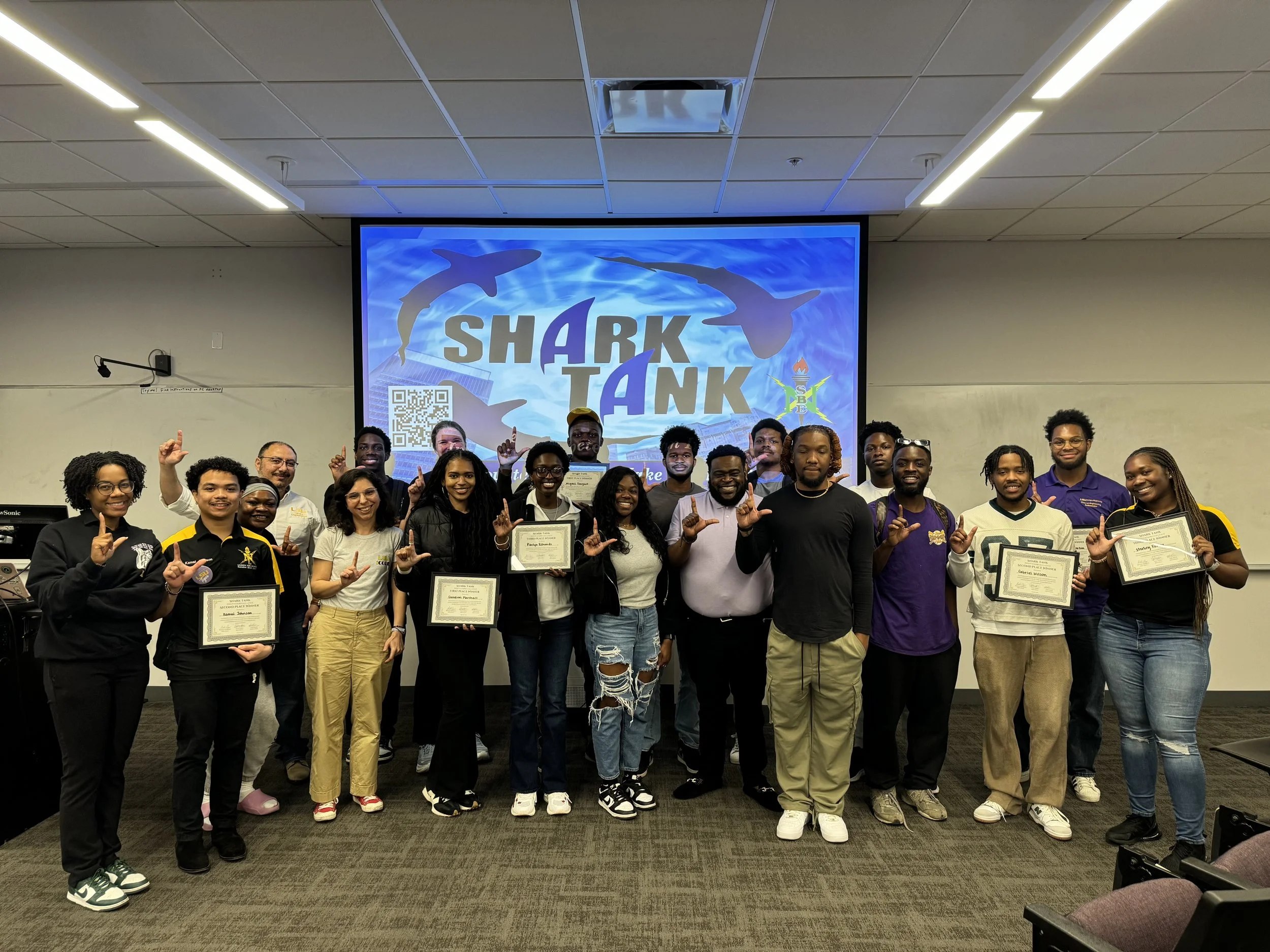 Group of diverse people holding certificates during a Shark Tank event in a conference room, with a large screen displaying the Shark Tank logo behind them.