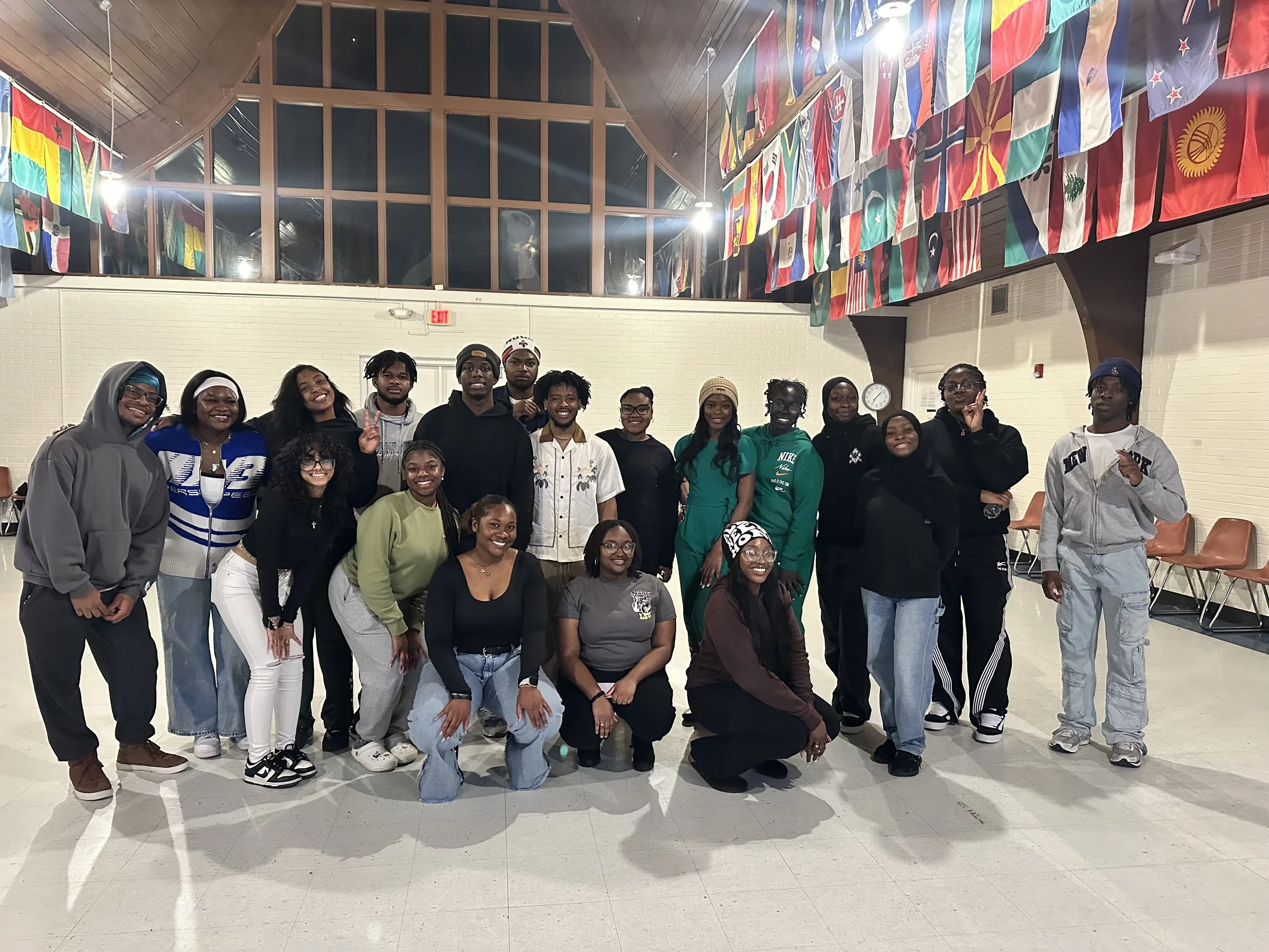 Group of diverse people posing together indoors, with international flags hanging from the ceiling.