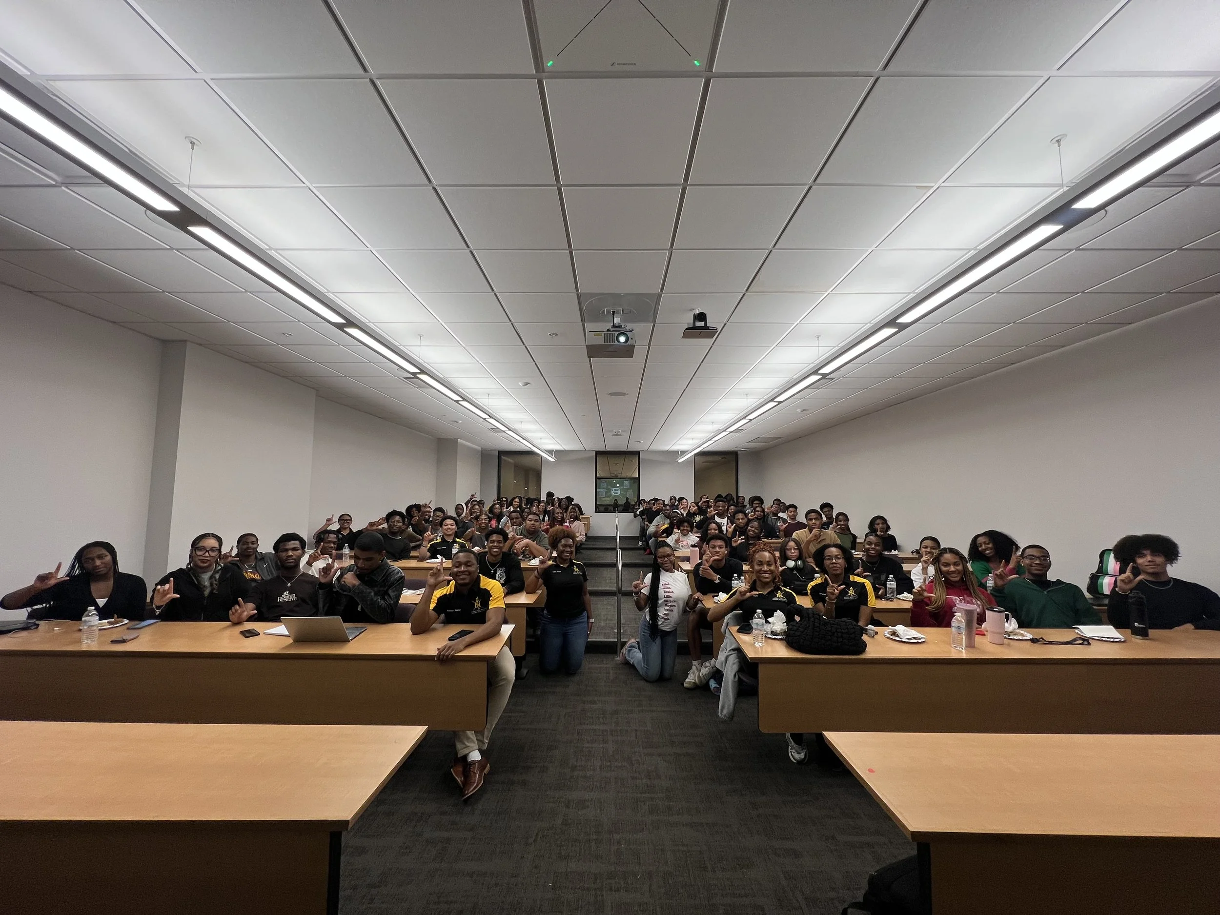 A large group of students sitting at long tables in a classroom or lecture hall, facing the camera, many making hand gestures, with some using laptops and water bottles, in a brightly lit room with white walls and a projector on the ceiling.