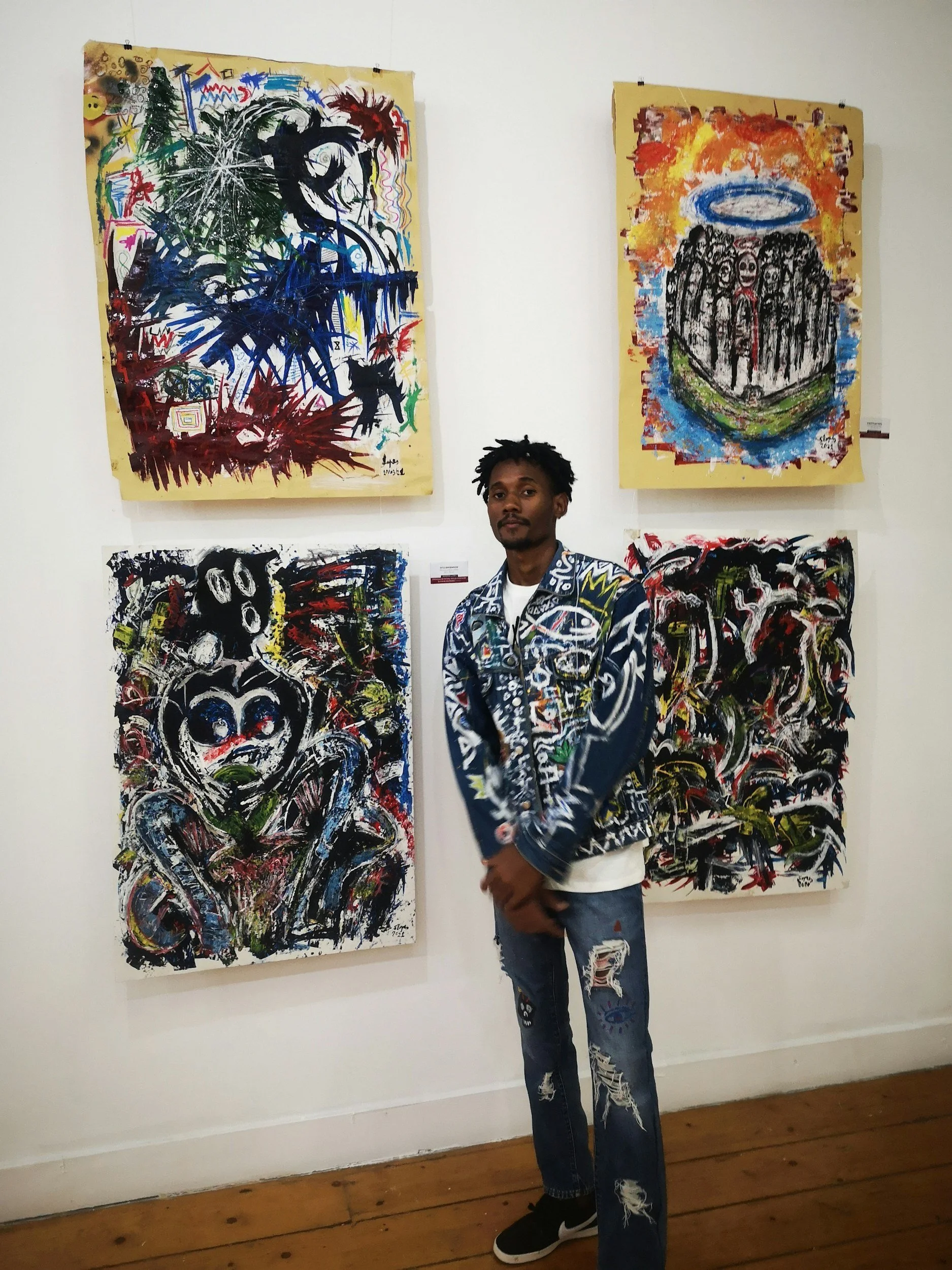 アートギャラリーの展示室にて、男性がサングラスをかけてポーズをとりながら、抽象画の前に立っている。彼は黒のスニーカーとジーンズ、ジャケットを着ている。背景には複数の鮮やかな色彩と抽象的な絵画が飾られている。