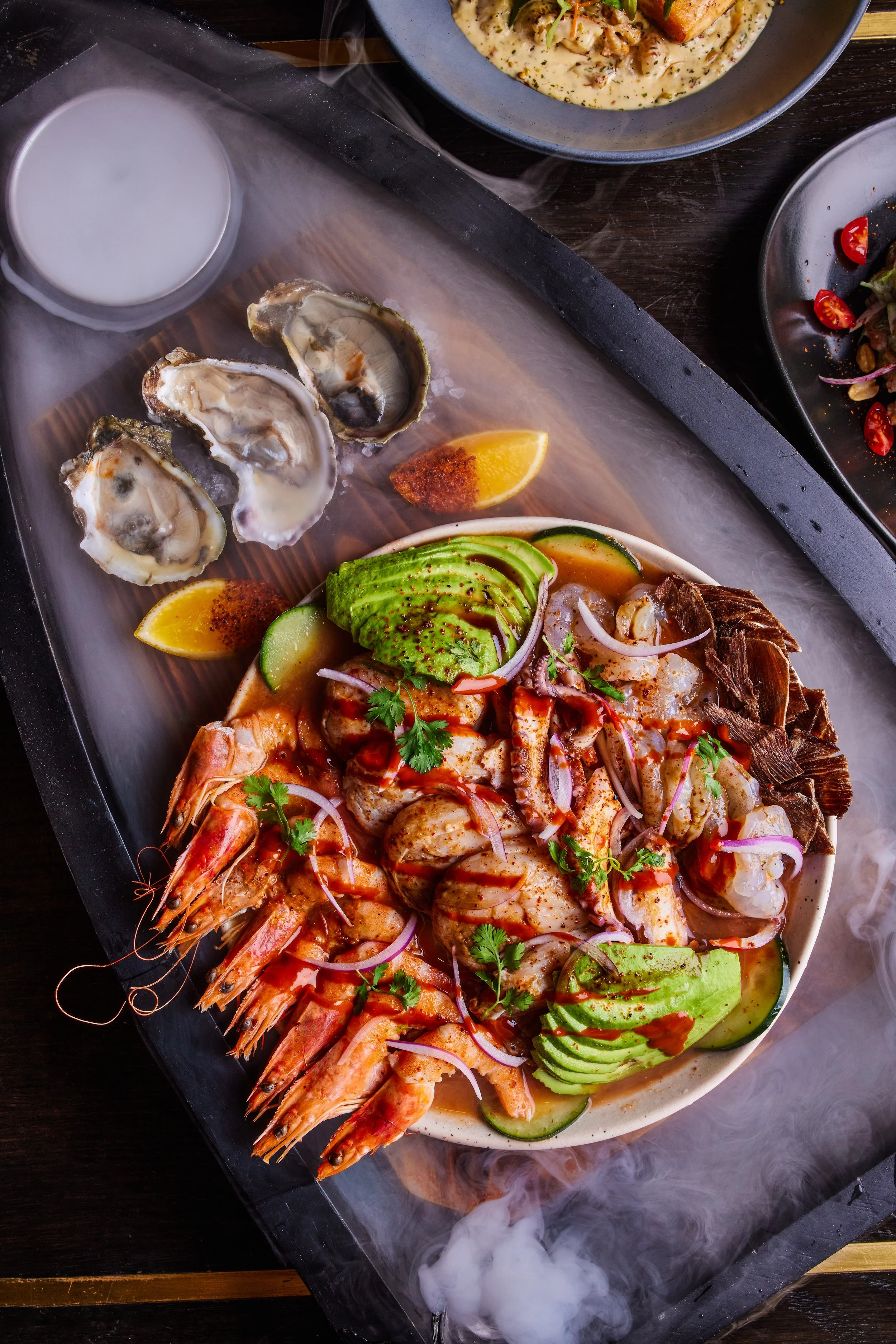 A tray with a seafood platter including oysters on the shell, shrimp, octopus, and sliced avocado, garnished with cilantro and red onions, served with lemon wedges and a small container of sauce or ice; the tray is emitting smoke or vapor.