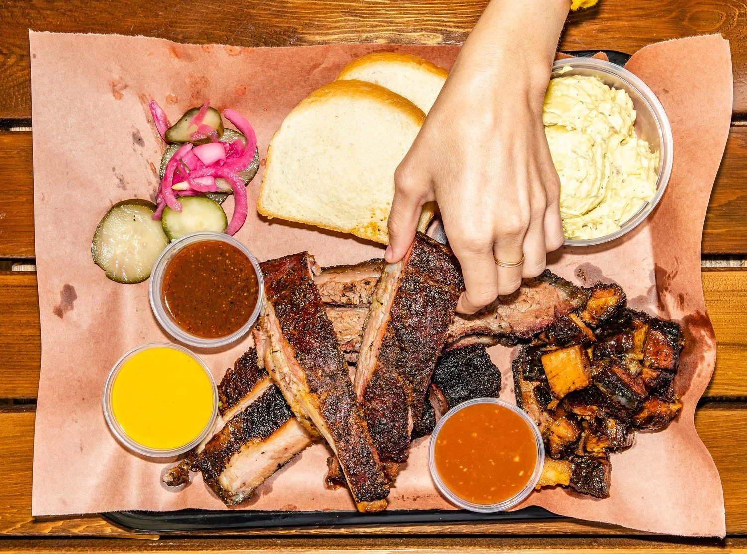 Barbecue ribs with slices of bread, pickles and onions, four dipping sauces, and a container of potato salad on a tray lined with pink paper.
