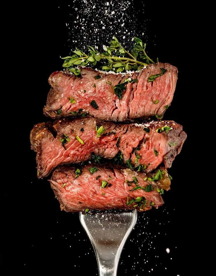 Three slices of cooked steak garnished with chopped herbs and salt on a fork, with some salt sprinkled above against a black background.