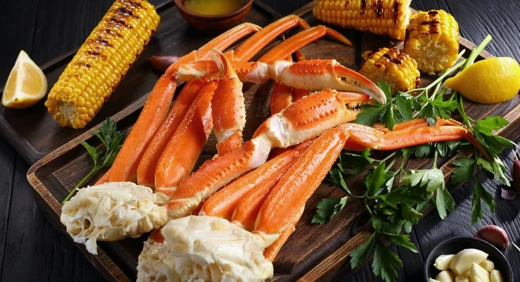 Crab legs with grilled corn, lemon wedge, parsley, and garlic on a wooden serving board.