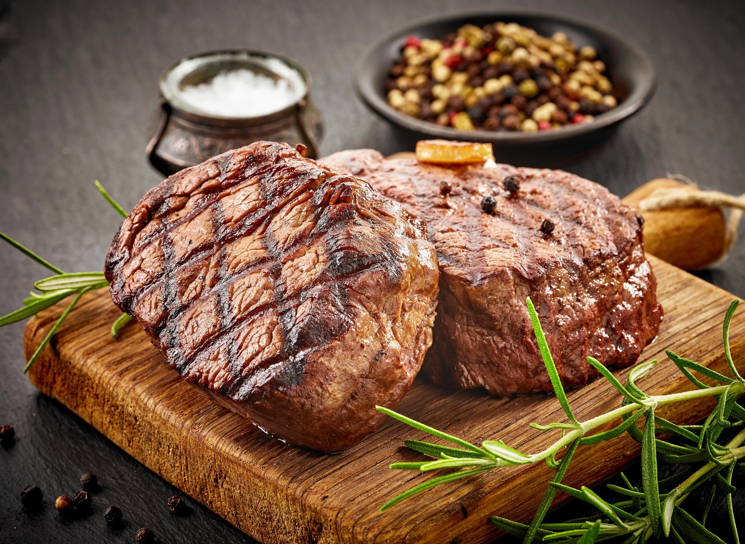 Two grilled cuts of steak on a wooden cutting board, garnished with rosemary sprigs, with a bowl of mixed peppercorns and a small container of salt in the background.