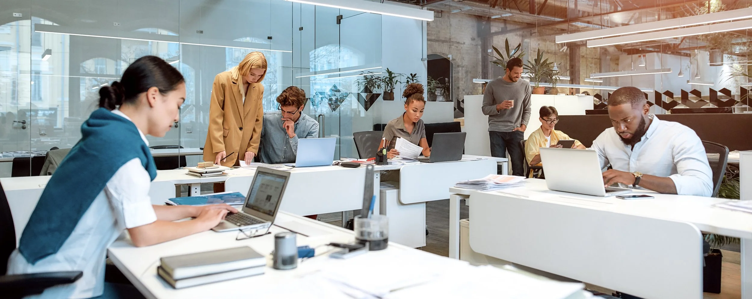 A diverse group of people working in an open-plan office with glass walls, laptops, and various office supplies, some standing and some seated at desks.