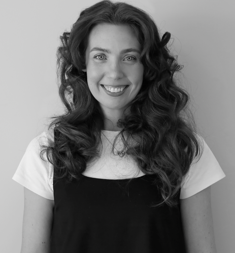 Black and white photo of a woman with long, curly hair smiling at the camera. She is wearing a short-sleeved top with a necklace. Graphic Designer Merridy Love.