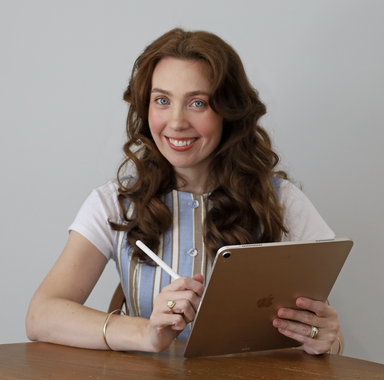 A woman with long, curly red hair and blue eyes using an iPad with an Apple Pencil, smiling at the camera, seated at a wooden table against a plain background. Graphic Designer Merridy Love.