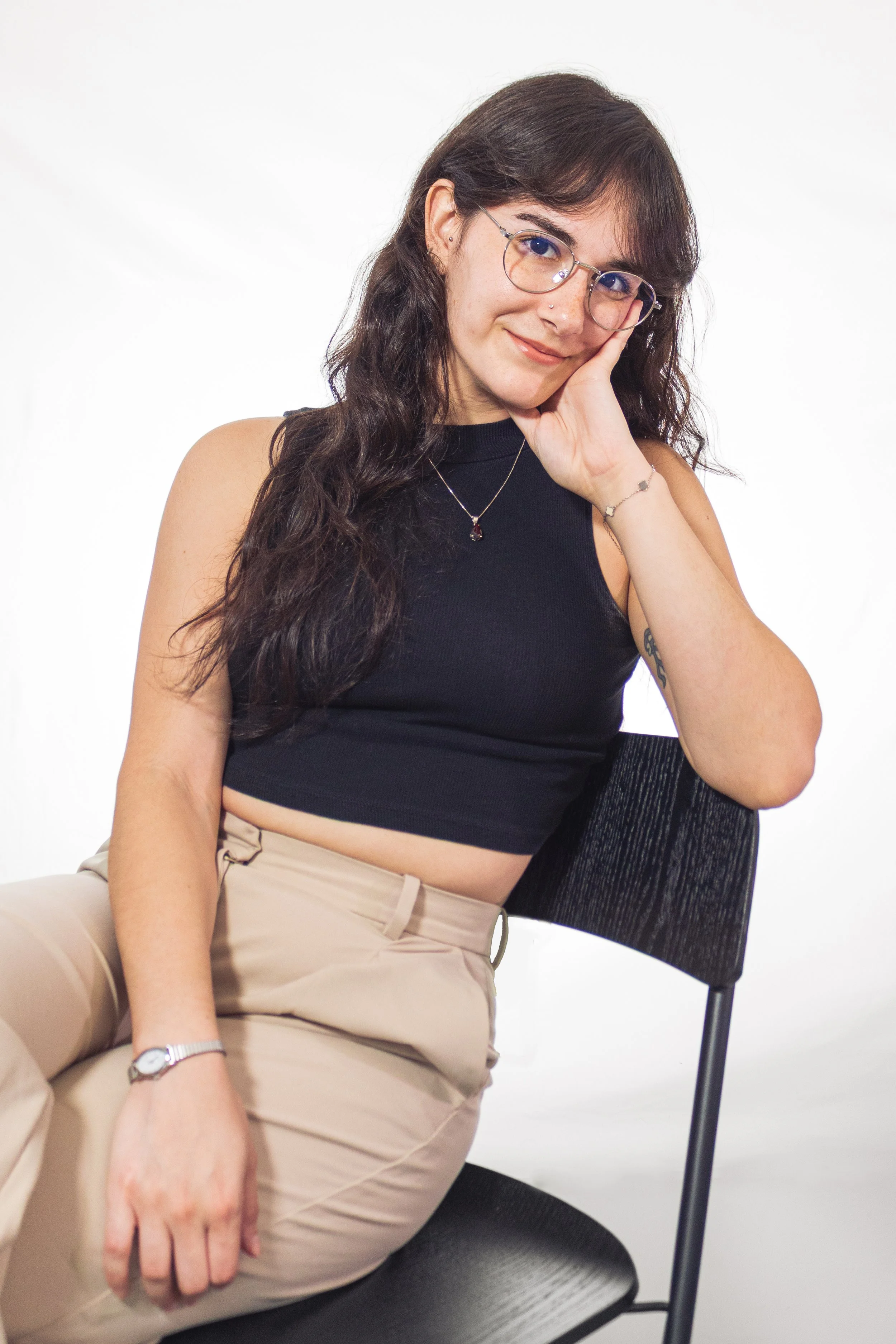 A young woman with long, wavy dark hair and glasses sitting on a black chair against a plain white background, smiling gently and resting her head on her hand.
