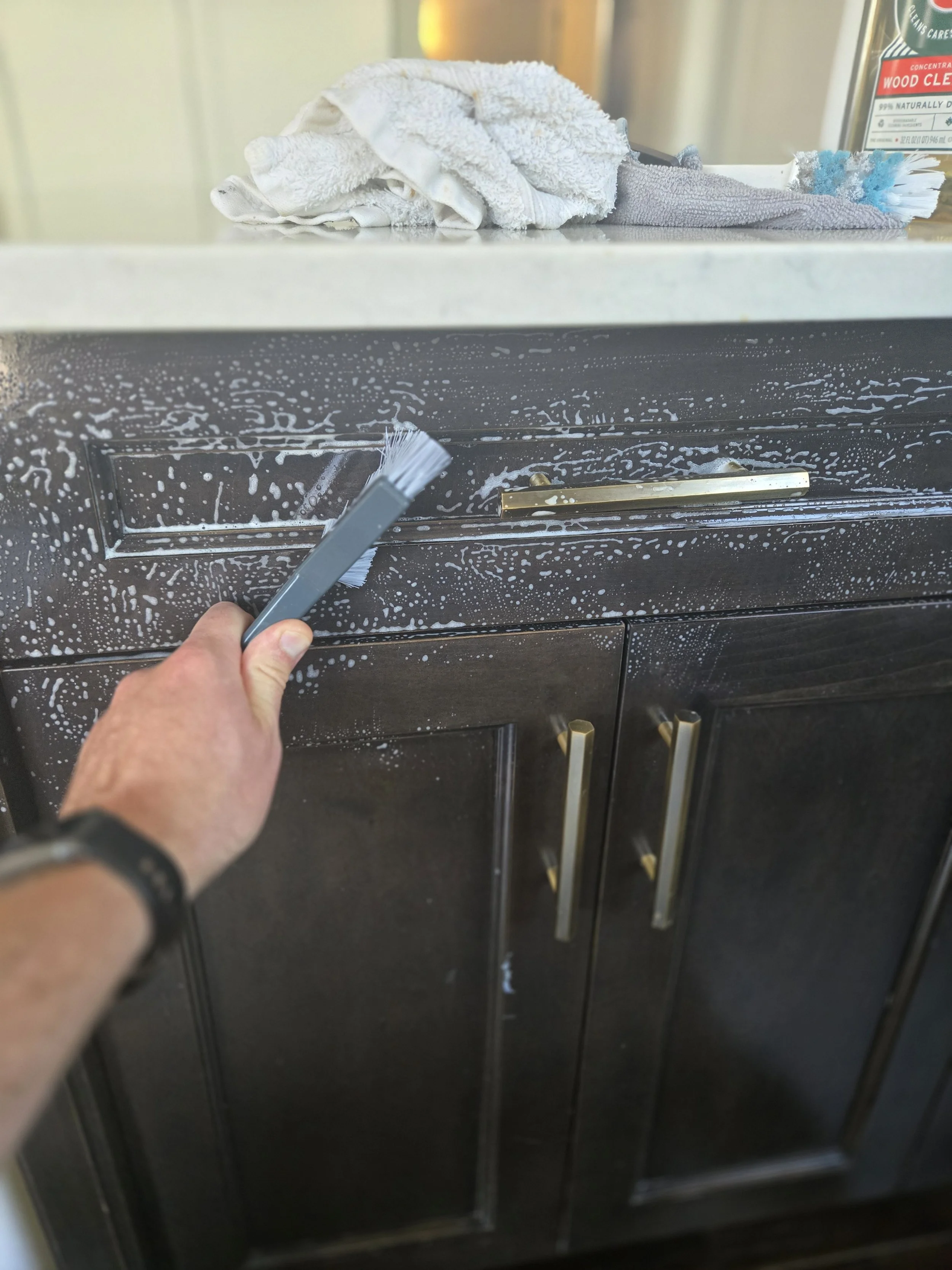 Person cleaning black kitchen cabinets with a brush, soap suds visible on surface, countertop with towels and spray bottle in the background.