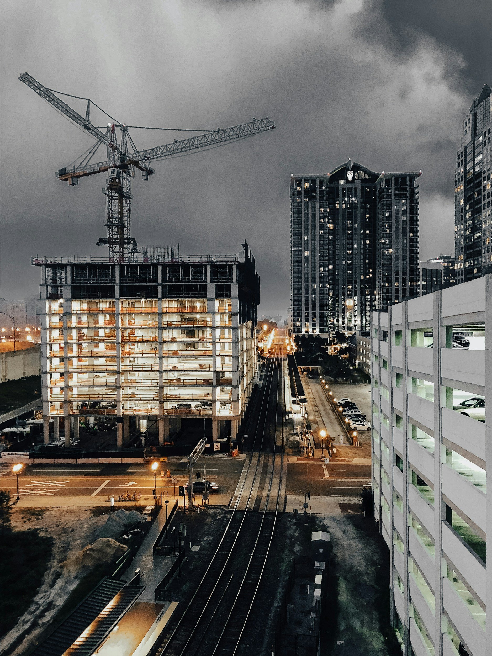 Nighttime urban construction scene with a tower crane above a partially built mid-rise structure, rail tracks running through the center, and high-rise buildings under dark storm clouds.