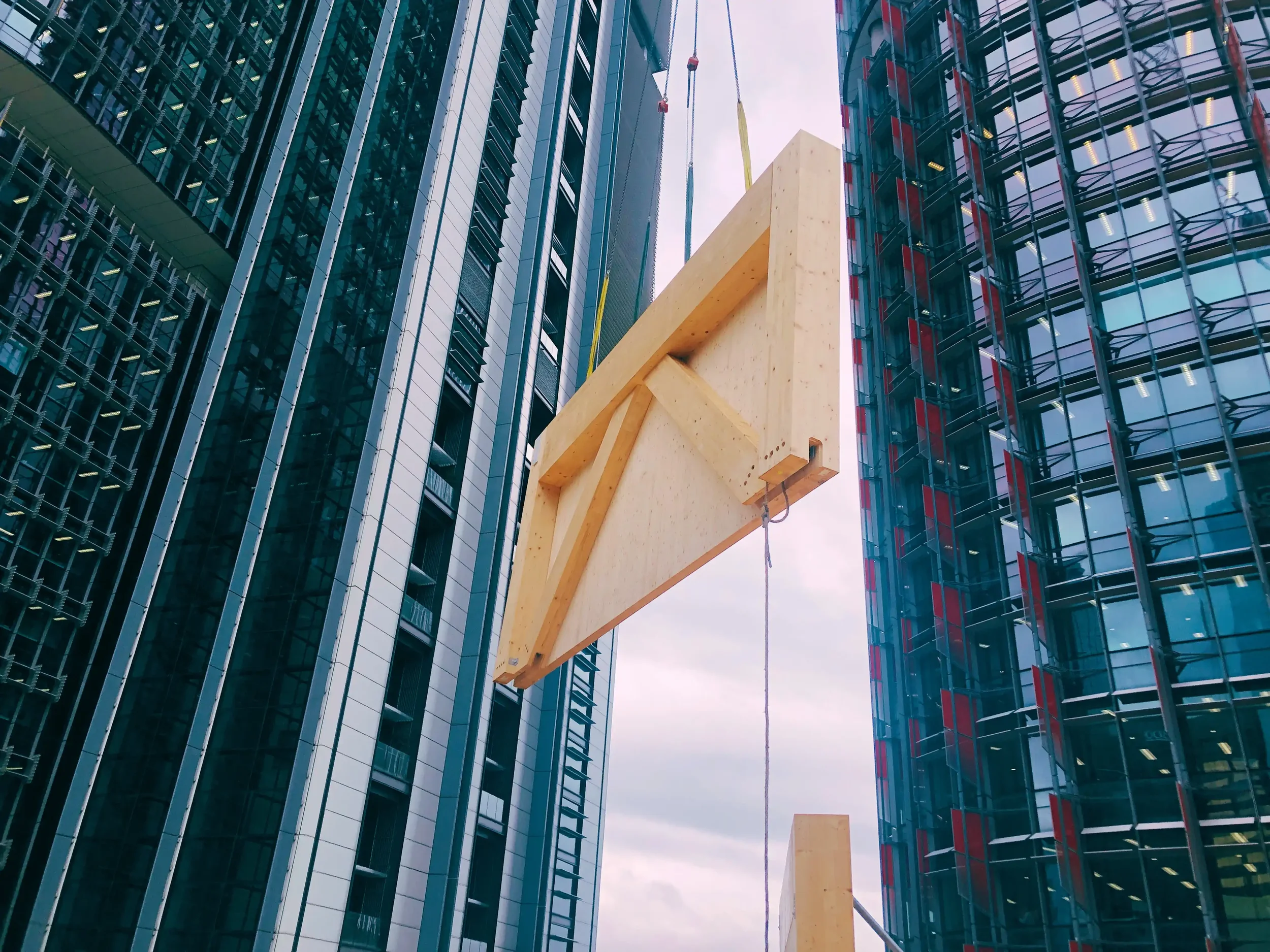 Cross-laminated timber (CLT) panel being lifted by crane between high-rise buildings, demonstrating prefabricated mass timber construction and modern methods of construction for urban development
