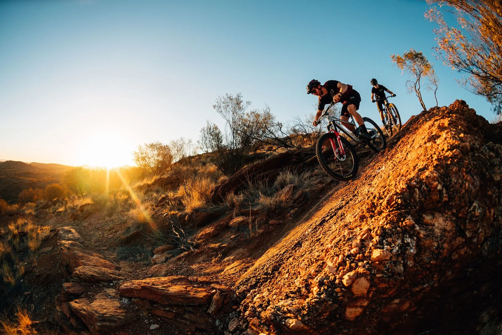 Two mountain bikers riding downhill on a rugged trail during sunset with trees and hills in the background.