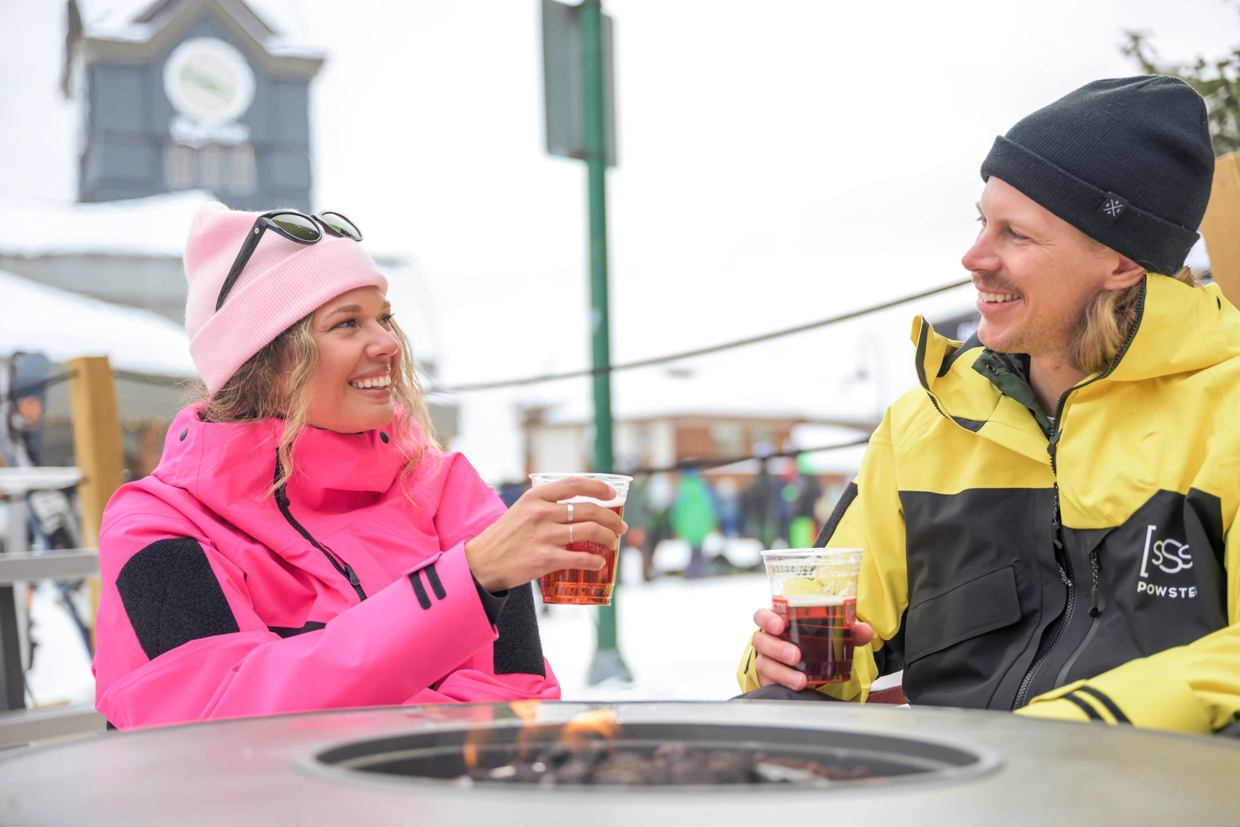 A woman and a man sitting outside in winter, smiling and holding cups of beer, dressed in bright outdoor winter jackets and hats, with a snowy background and a fire pit in front of them.