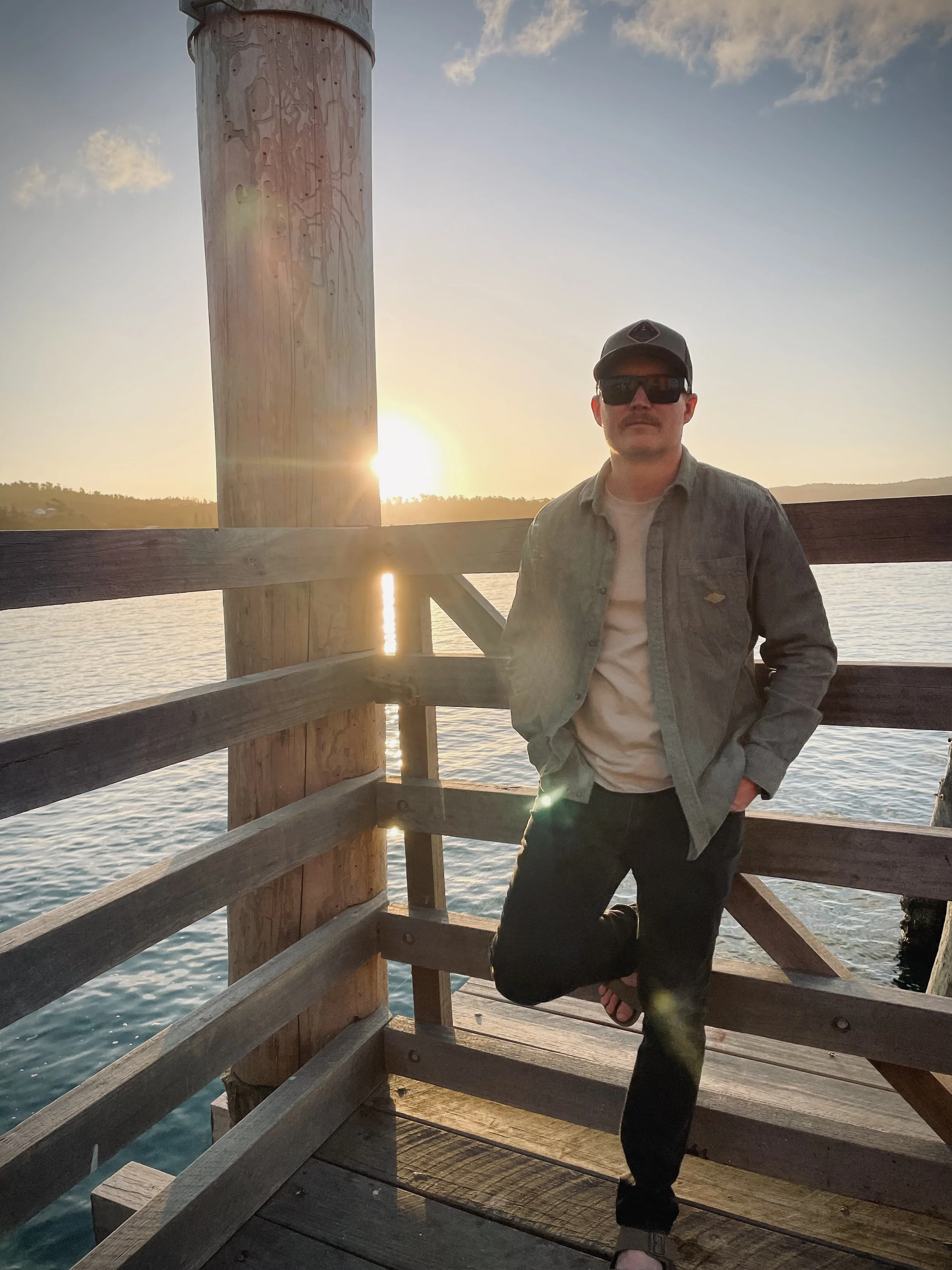 A man standing on a wooden pier at sunset, wearing a denim jacket, sunglasses, a cap, and flip-flops, with water and a distant shoreline in the background.